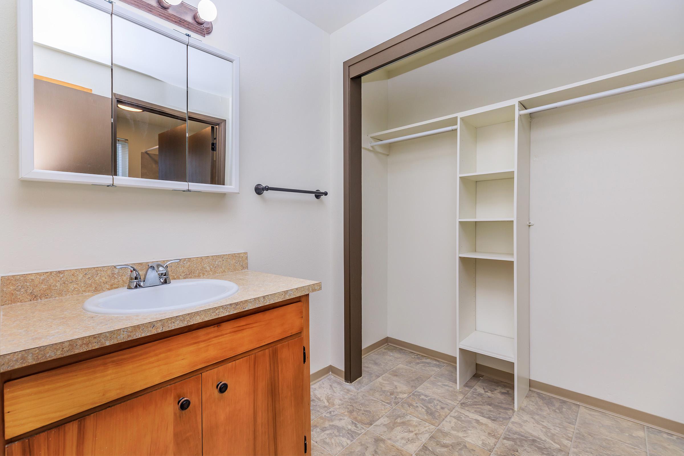 A spacious bathroom featuring a wooden vanity with a sink, a mirror above it, and a towel rack on the wall. Adjacent to the vanity is a well-organized closet space with shelving and an open hanging area, all set against a neutral-colored wall and tiled floor.