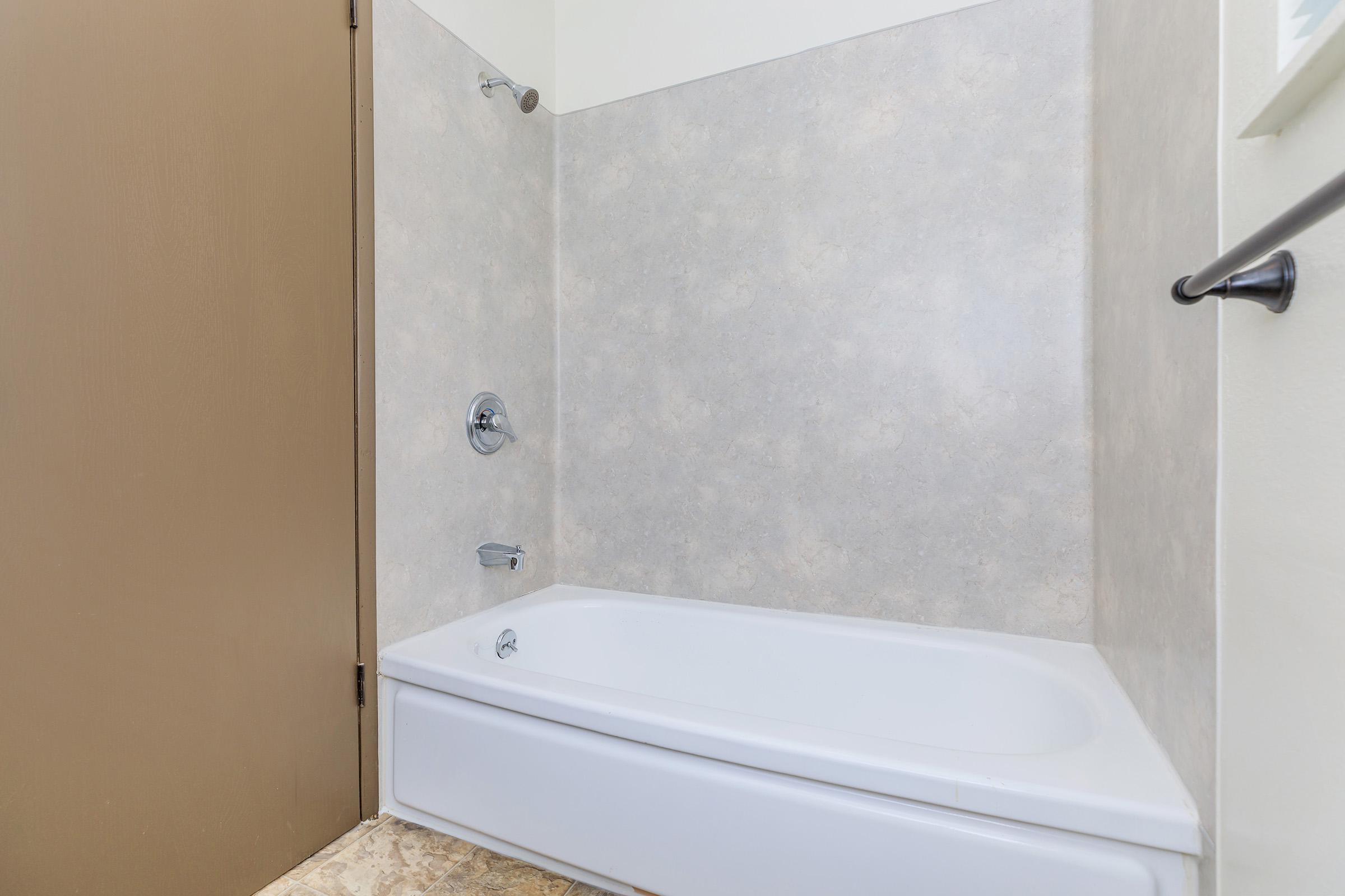 A clean, empty bathtub with a showerhead mounted on the wall. The surrounding walls are a light, textured material, and there’s a dark brown door nearby. The floor features light-colored tiles, creating a simple and inviting bathroom space.