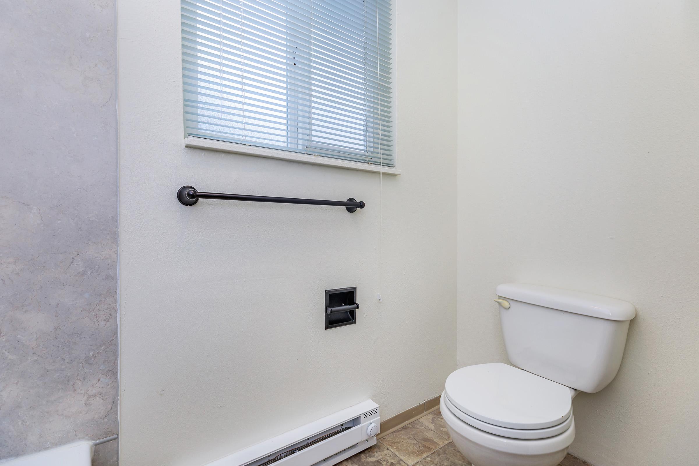 A clean, minimalist bathroom featuring a white toilet, a towel bar, and a small window with blinds. The walls are a light color, and the floor has a textured tile pattern. A heating unit is visible near the floor, and there is no additional decor, creating a simple, functional space.