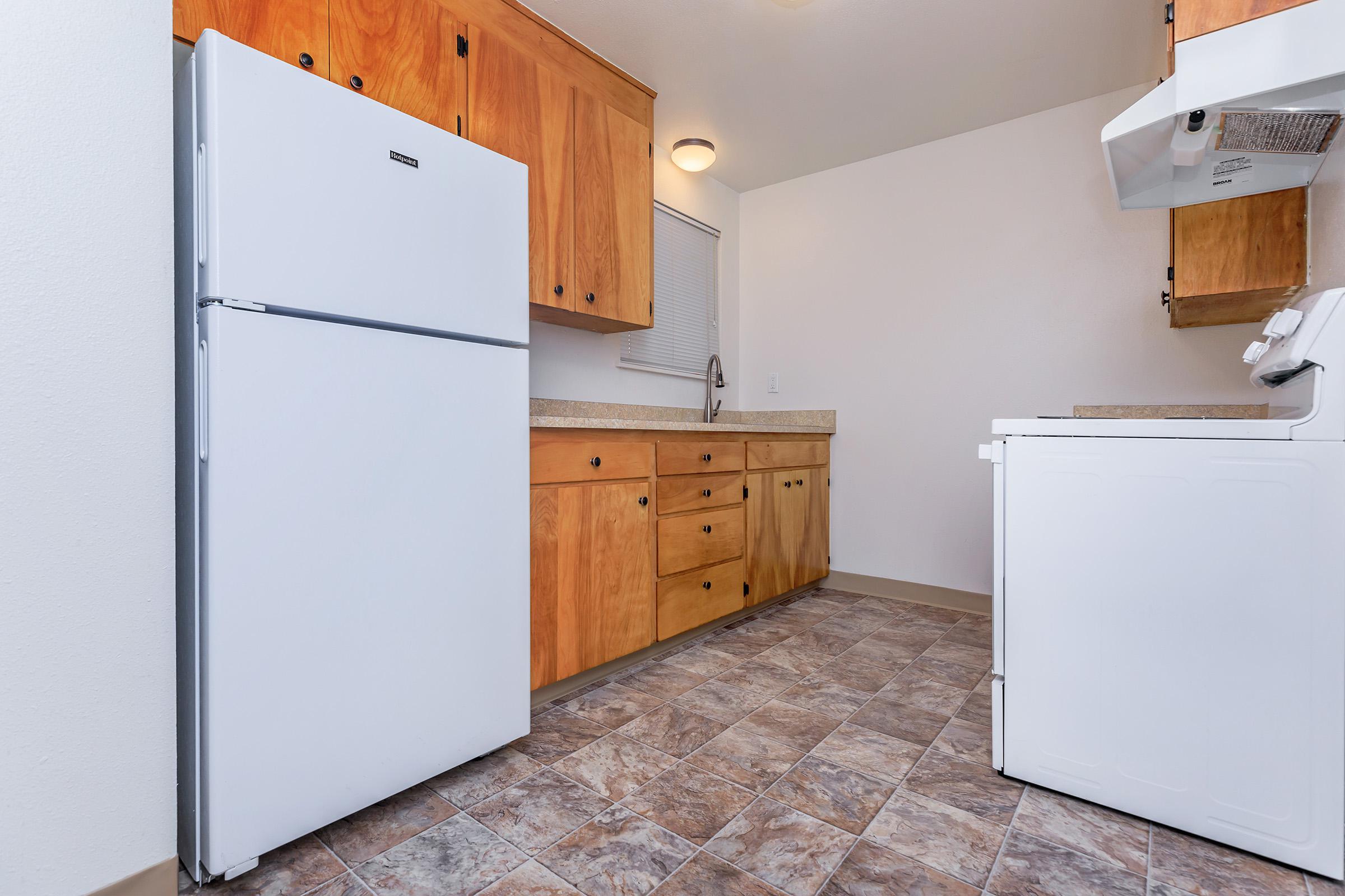 A view of a modern kitchen featuring a white refrigerator and a washing machine. The kitchen has wooden cabinets, a countertop, and tiled flooring. The walls are painted a light color, and there is a small window for natural light. Overall, it presents a clean and organized space.