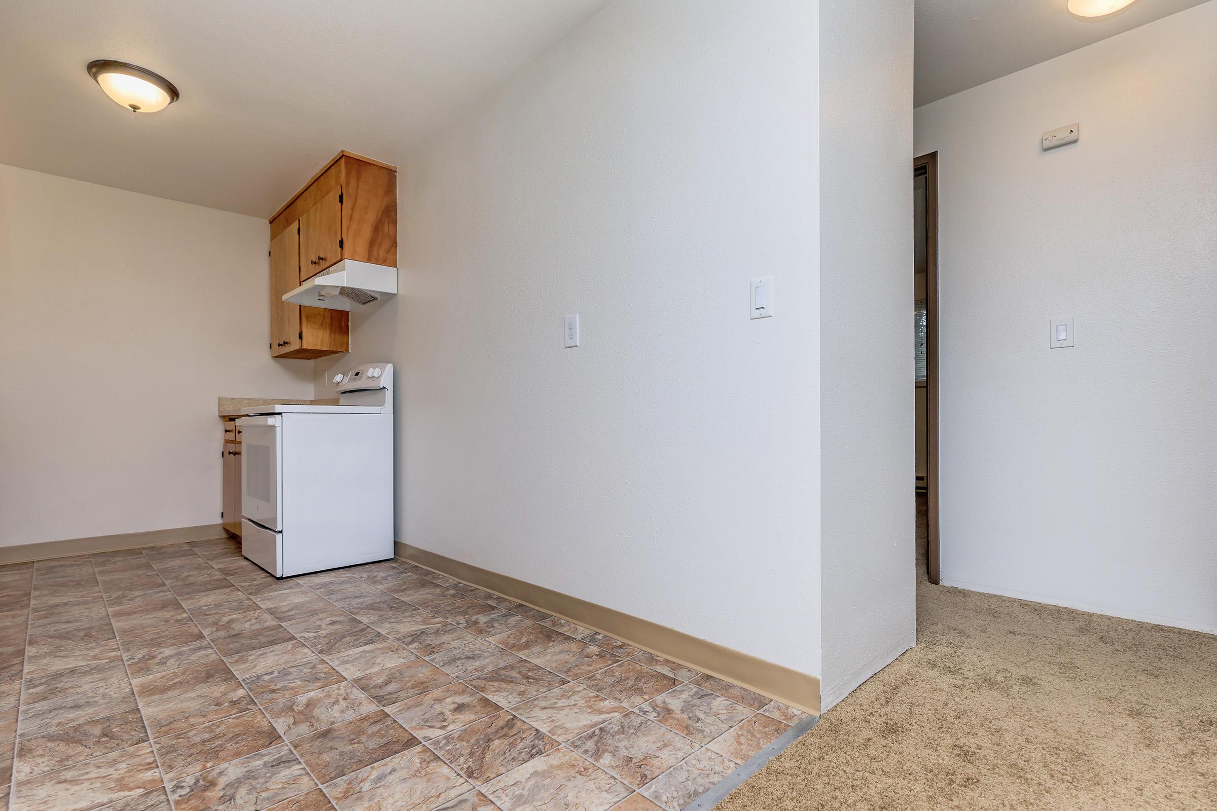 A small, empty kitchen area featuring light-colored walls, a tiled floor, a white stove, and a kitchen cabinet. A doorway leads to another room, with a light fixture on the ceiling providing illumination. The overall ambiance is clean and simple, suitable for a compact living space.