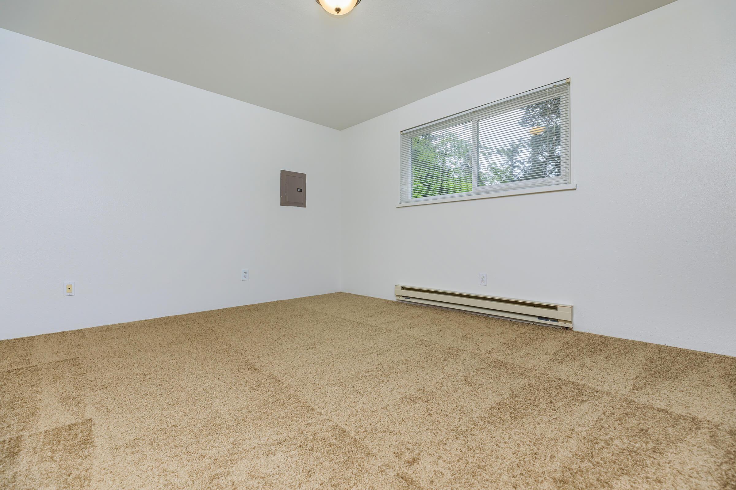A simple, empty room with beige carpet and white walls. A small window lets in natural light, and there is a utility panel on one wall. The overall atmosphere is clean and minimalistic, suitable for various uses such as a bedroom or office space.