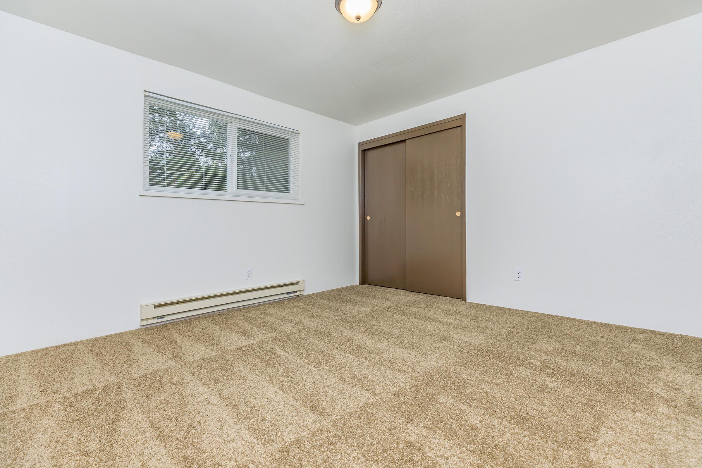A well-lit, empty room featuring light beige carpet flooring, a window with blinds, and a brown closet door. The walls are painted white, and there is a ceiling light fixture. The room appears spacious and clean, suitable for various uses.