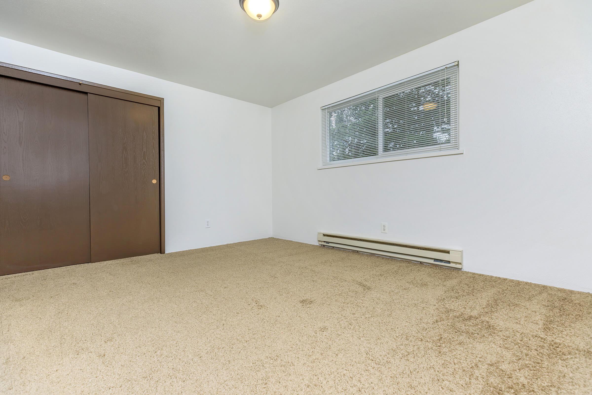 Spacious empty room with beige carpet, featuring a single window with blinds and a ceiling light. There are brown double doors on one wall, suggesting storage space. The walls are painted white, and the overall atmosphere is bright and neutral, suitable for various uses.