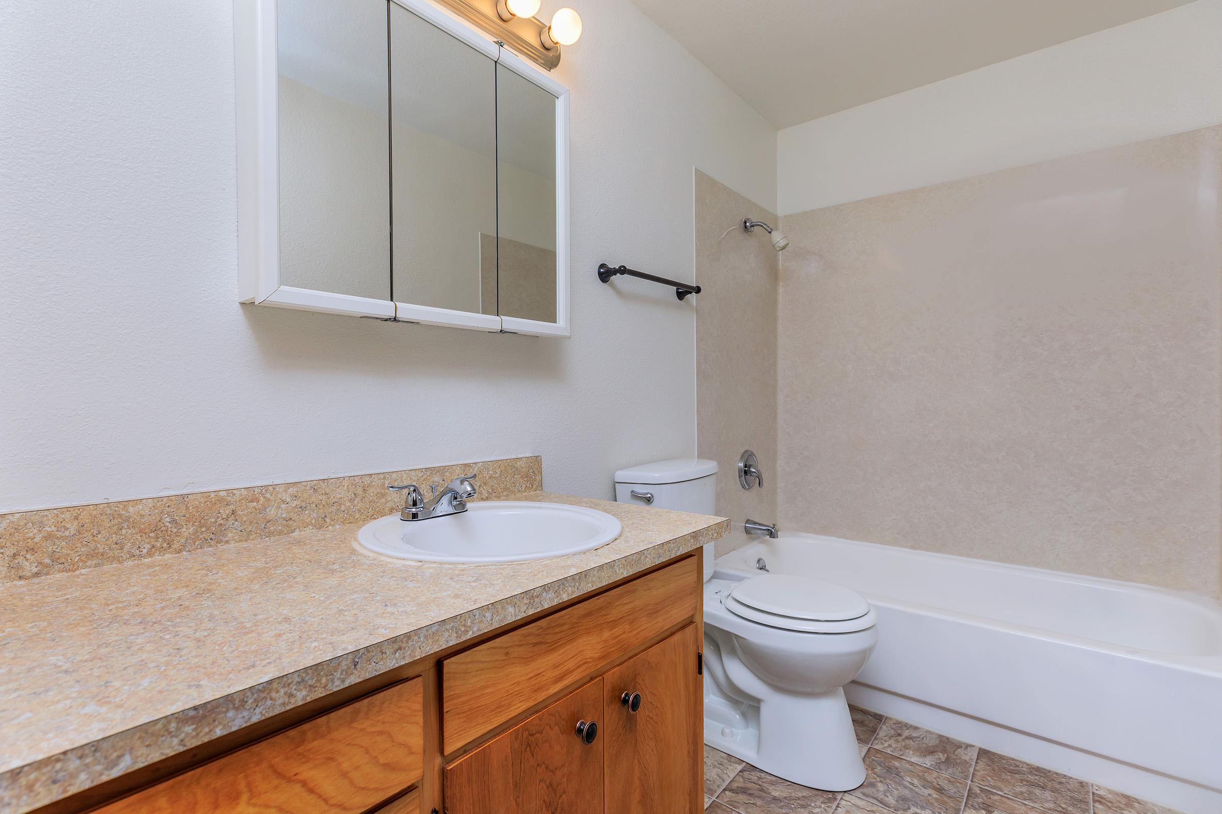 A clean and modern bathroom featuring a light-colored wall, a wooden vanity with a sink, a mirror above the sink, and a bathtub with a shower. The toilet is adjacent to the bathtub, and the floor is tiled with a brown pattern. Natural light illuminates the space, creating a bright atmosphere.