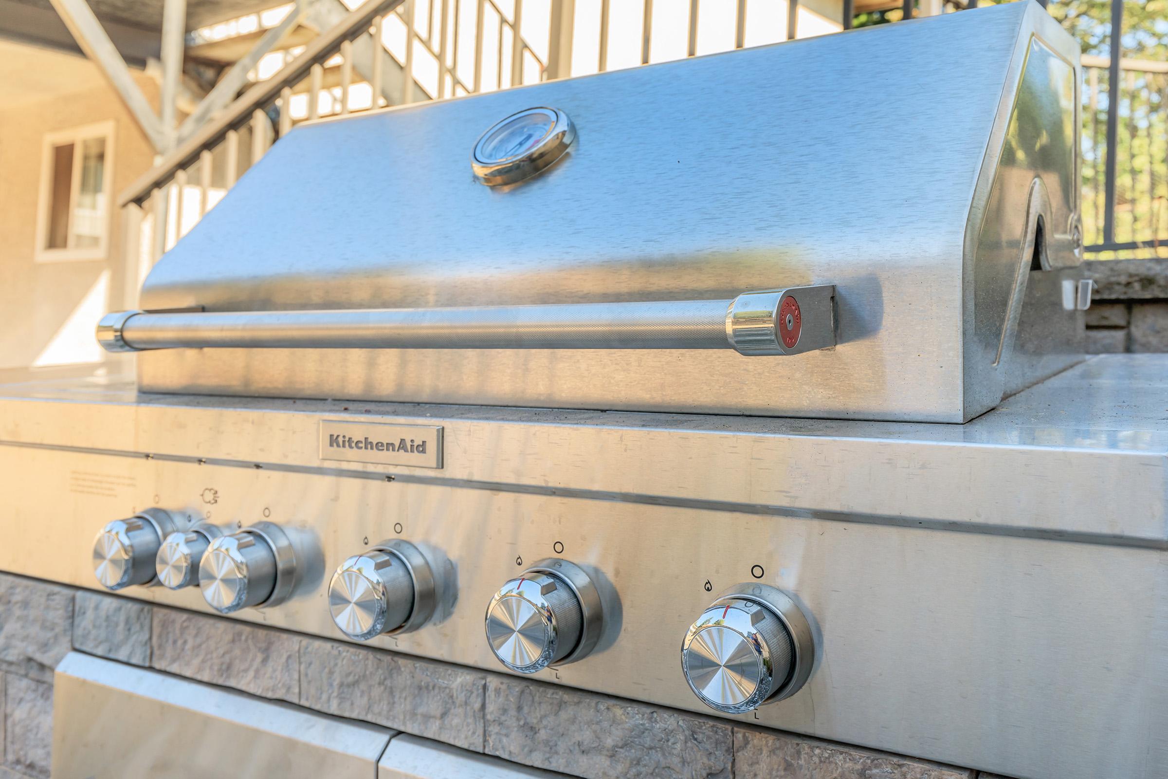 A stainless steel KitchenAid grill with a closed lid and five control knobs at the front. The grill is positioned on a stone base, with a background of a house and stairs leading up. The overall setting suggests an outdoor cooking area.