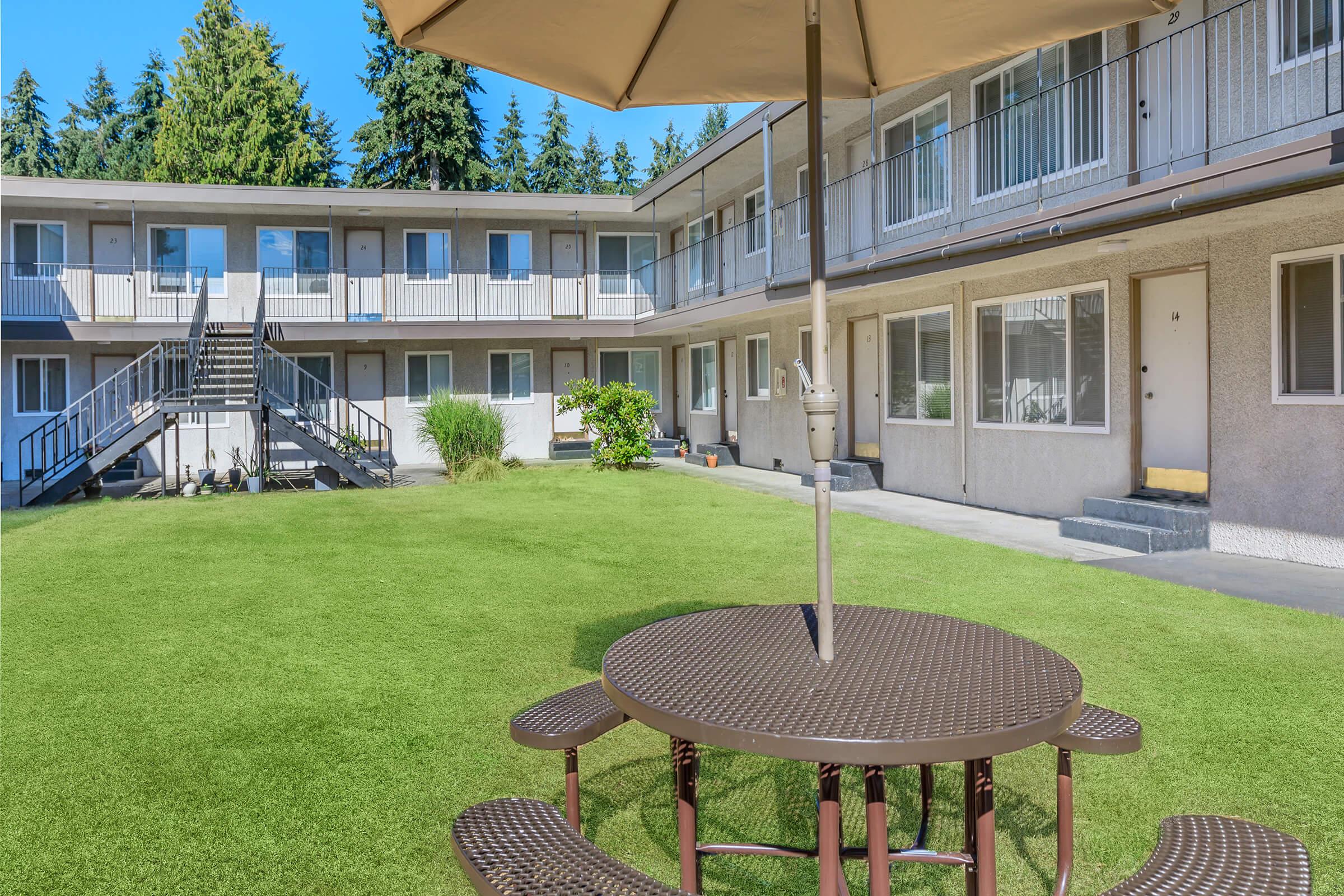 A well-maintained courtyard featuring a round picnic table with an umbrella, surrounded by lush green grass. In the background, there are two levels of a building with balconies and steps leading up, set amidst tall trees. The area appears clean and inviting, suitable for relaxation or outdoor gatherings.