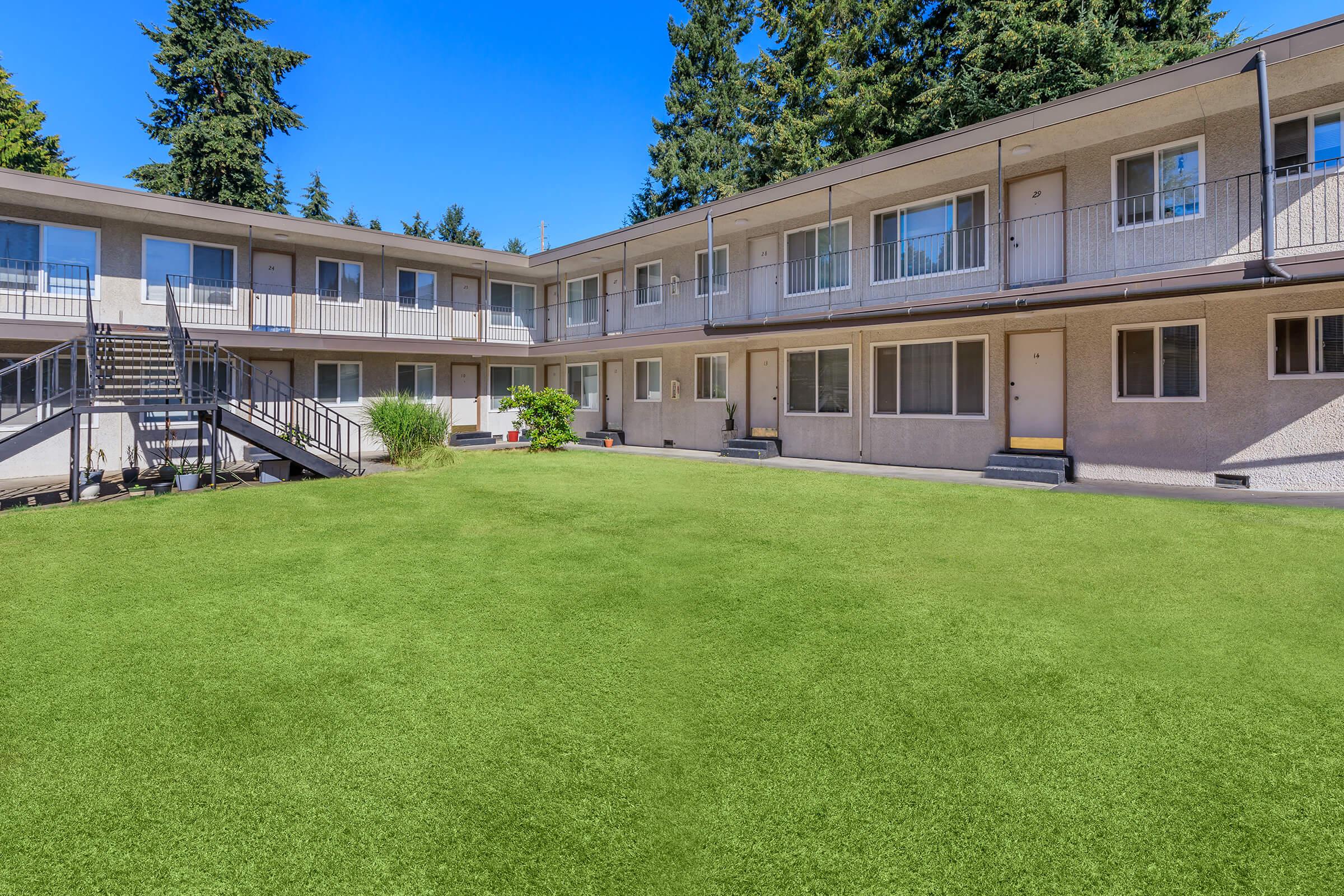 A view of a well-maintained outdoor area in front of a two-story apartment building. The building features several balconies and windows, surrounded by lush green grass and shrubs. Clear blue skies and tall trees provide a scenic backdrop.