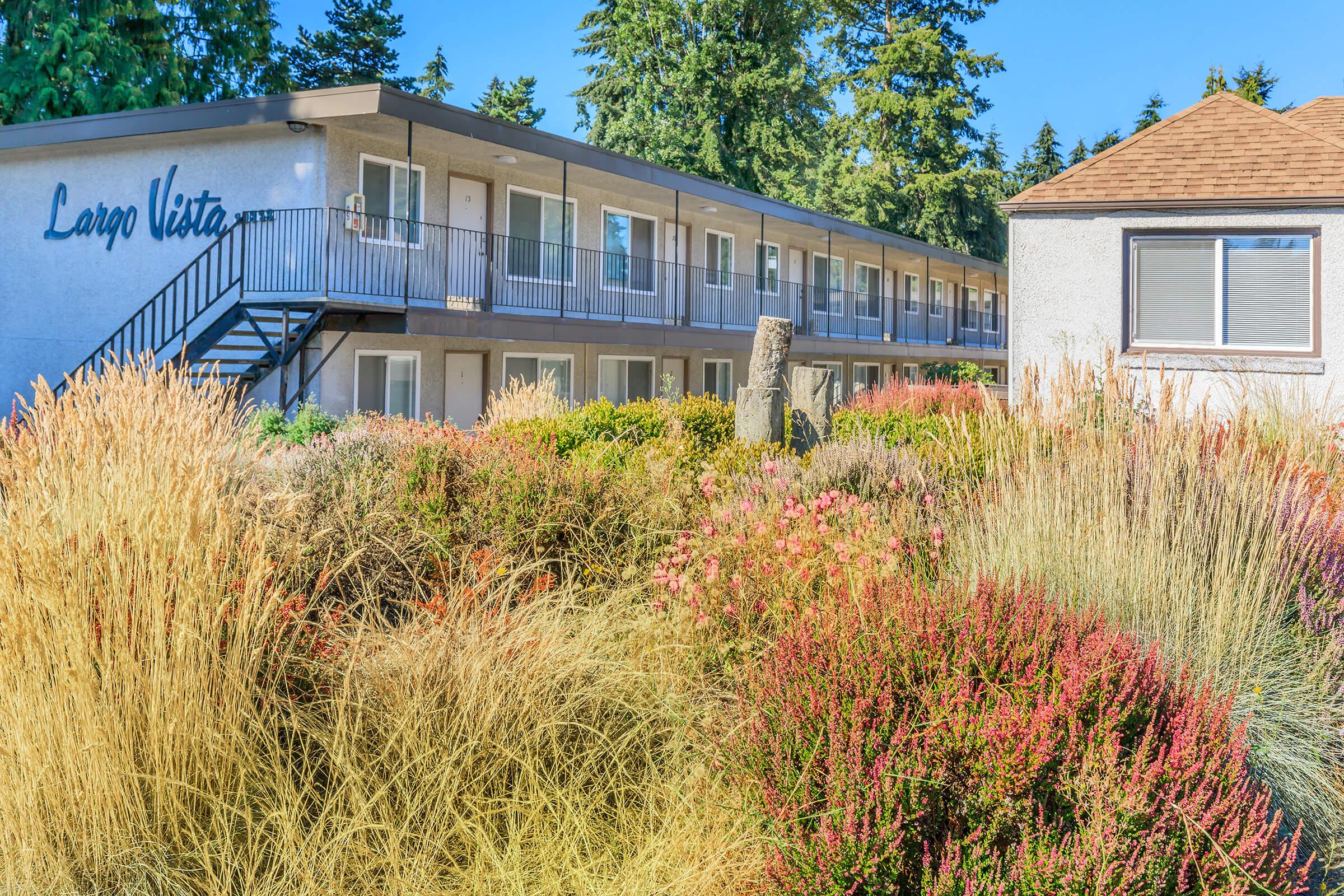 A well-maintained courtyard featuring colorful shrubs and tall grasses, with a multi-story building labeled "Largo Vista" in the background. The scene is surrounded by lush evergreen trees and a clear blue sky, creating a serene outdoor atmosphere.