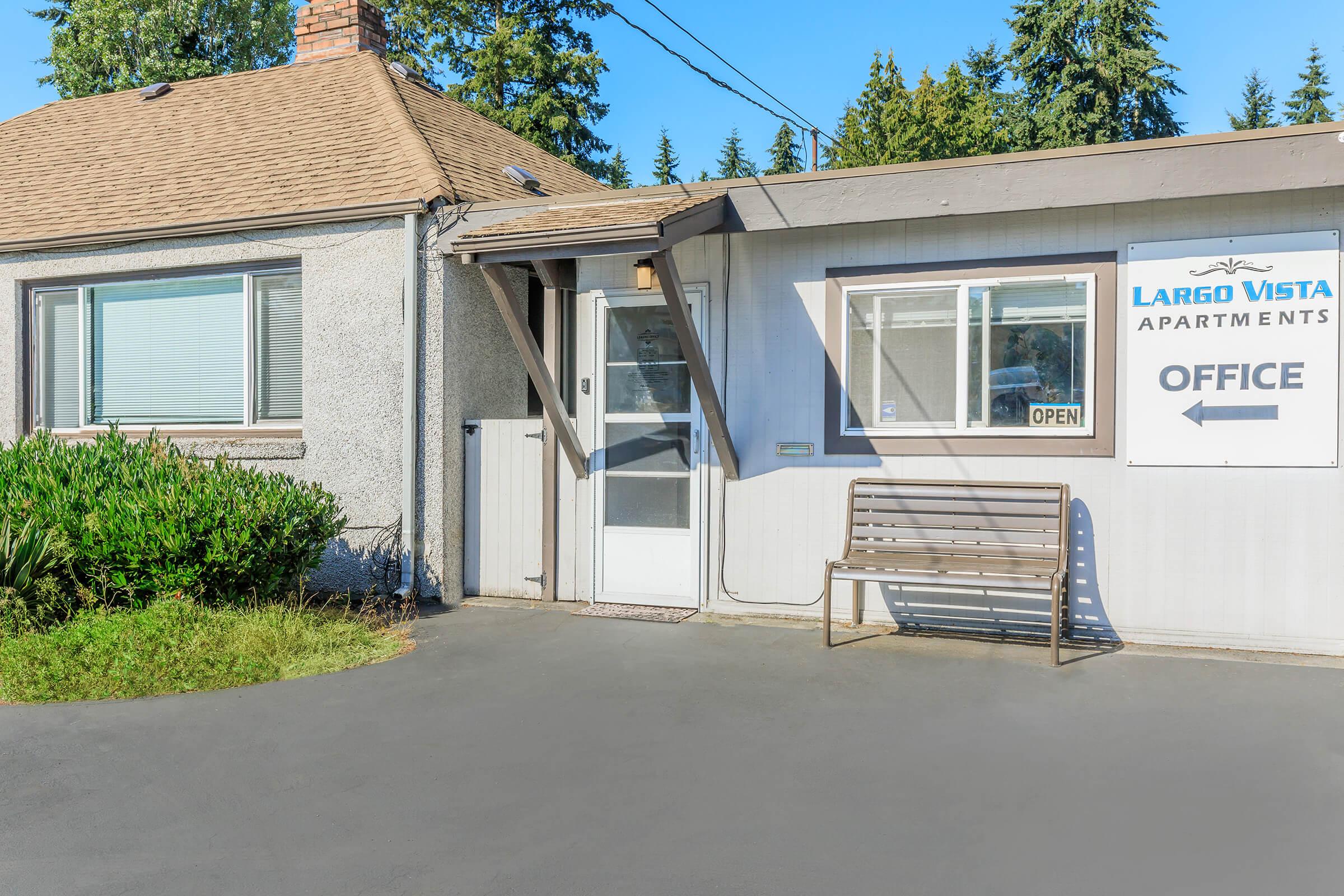 A small building with a light gray exterior and a brown shingled roof. The entrance features a glass door and a sign that reads "LARGO VISTA APARTMENTS - OFFICE." There’s a wooden bench in front, and well-maintained greenery surrounds the entrance. The sign indicates the office is open.