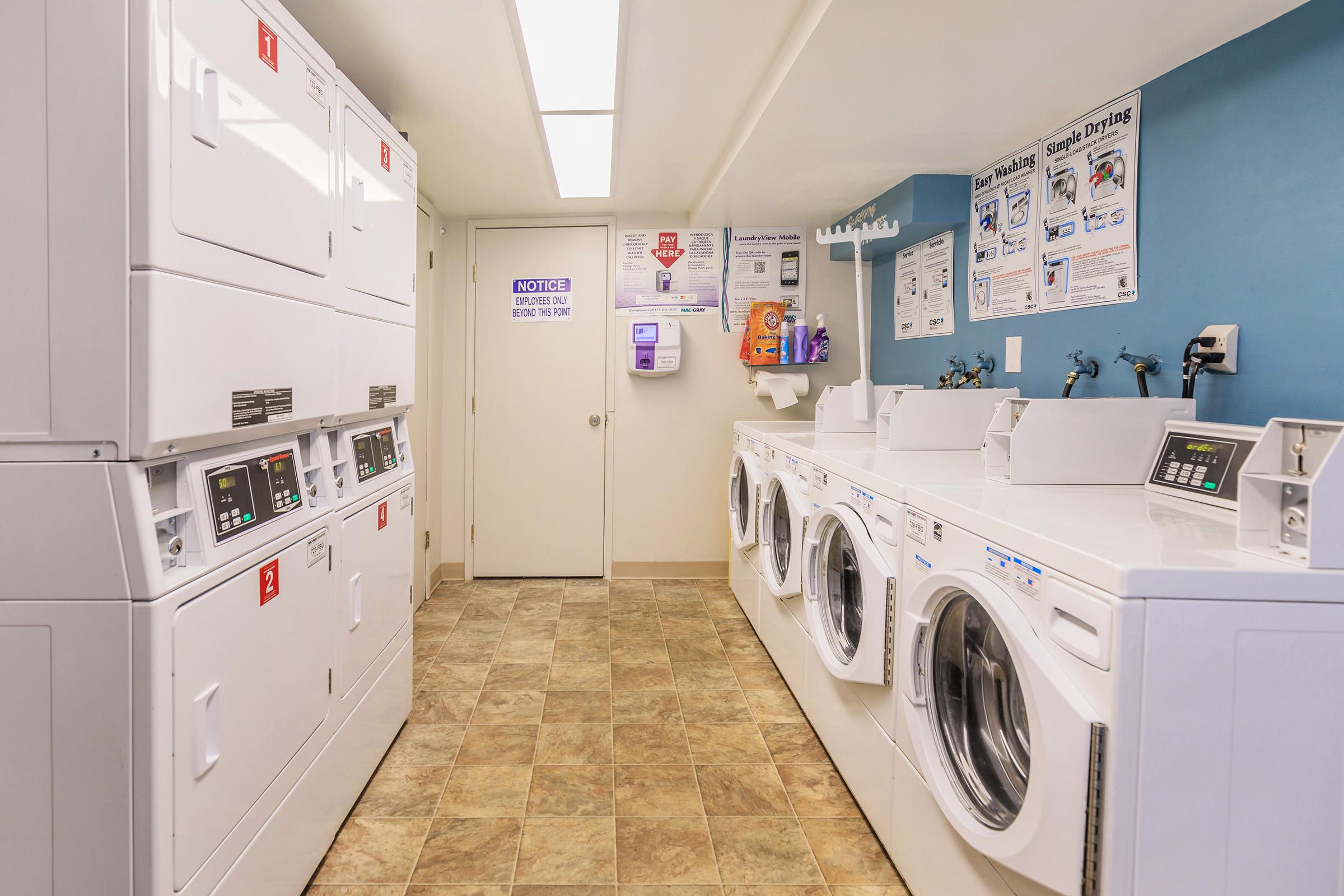 A clean and well-lit laundry room featuring several white washing machines and dryers lined up against the wall, with vending machines and signage on the walls. The floor is tiled in a beige pattern, and there is a small window allowing natural light to enter.