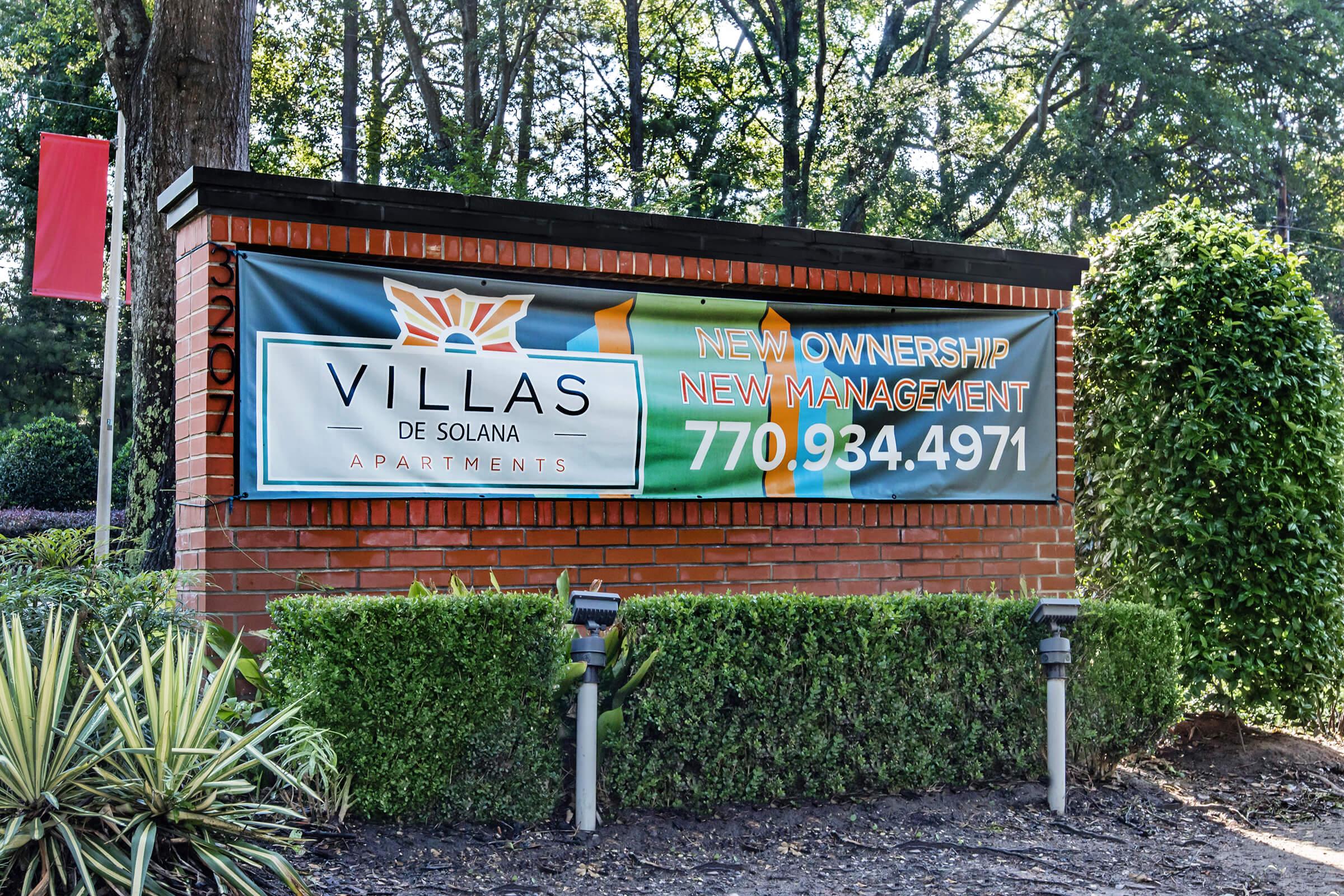 Sign for Villas de Solana Apartments announcing new ownership and management, featuring a colorful design with the text "Villas de Solana" prominently displayed. Contact phone number is listed: 770-934-4971. Surrounding greenery provides a decorative touch to the entrance.