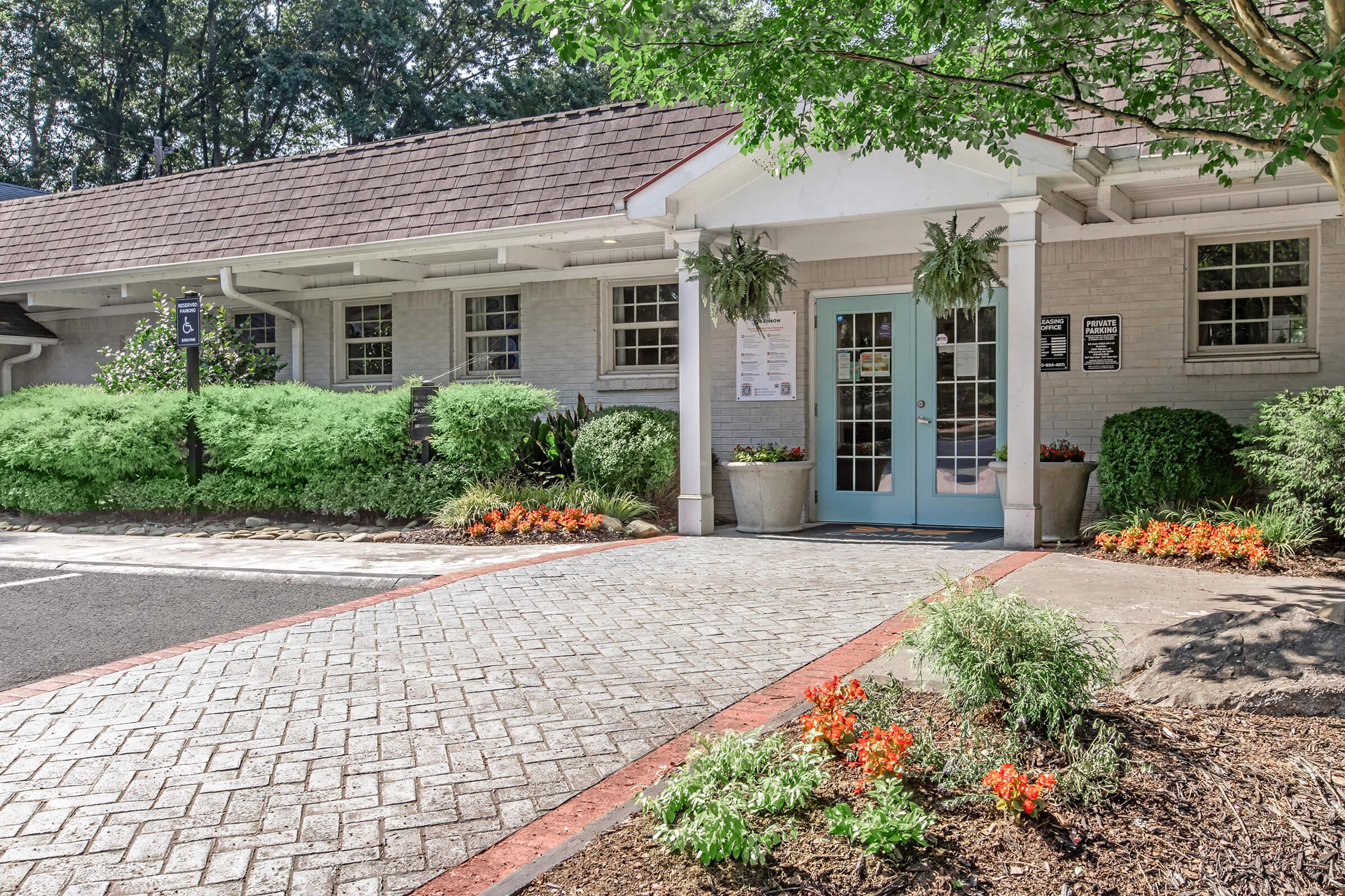 A welcoming entrance to a building with a light blue door framed by green plants and colorful flower beds. The pathway is paved with bricks, and there are lush green bushes on either side. Signs are posted next to the door. Trees provide shade in the background.