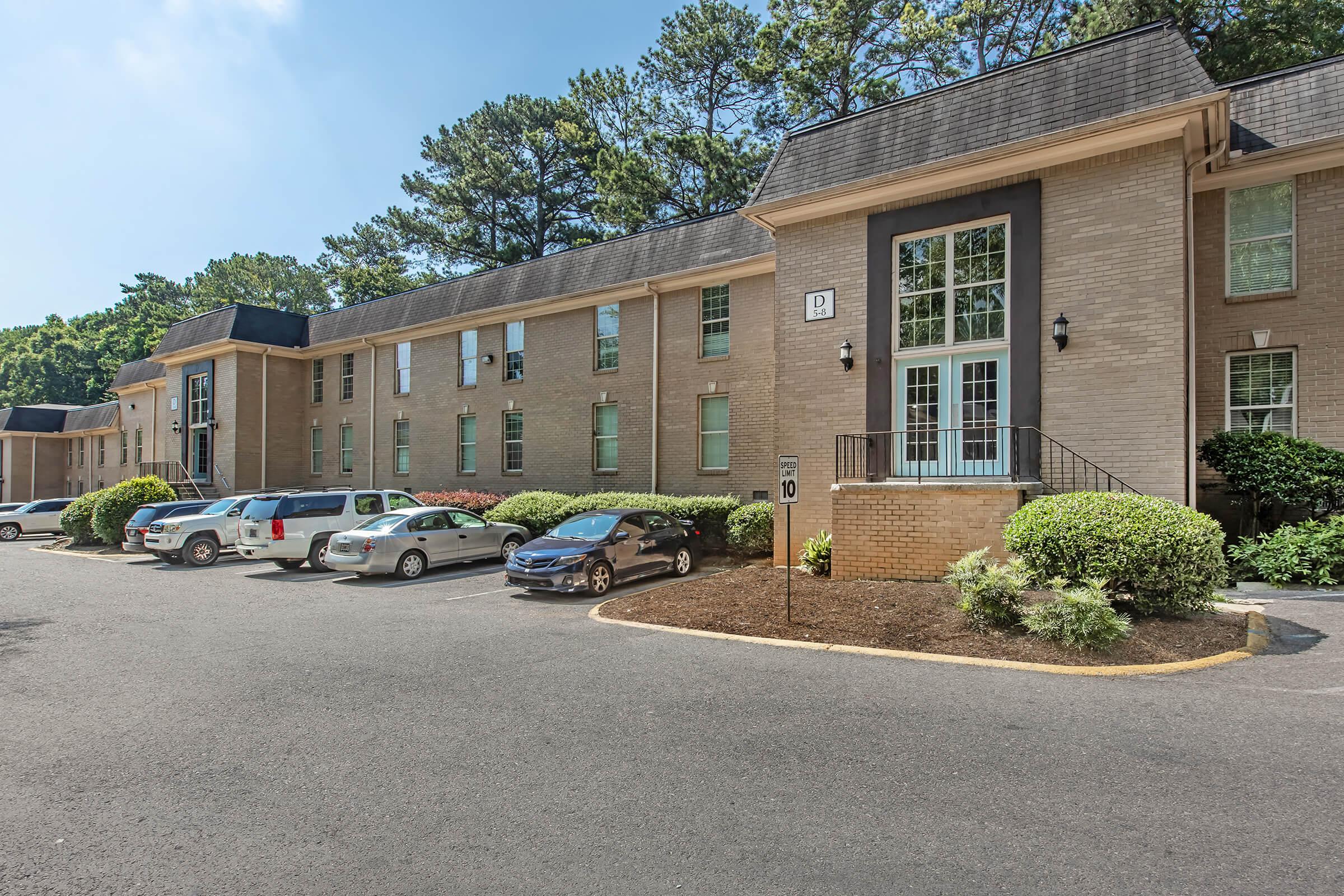 A beige apartment building with multiple windows and a central entrance surrounded by green trees and bushes. Several parked cars are visible in the foreground, with a clear blue sky above.