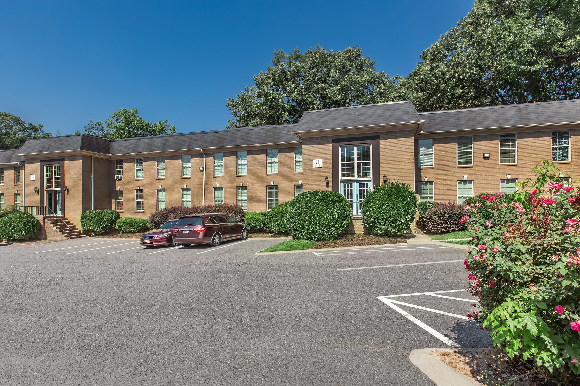 A two-story brick apartment building with several windows and shrubs in front. There's a parking lot with two parked cars and well-maintained landscaping featuring flowers. Trees provide shade in the background under a clear blue sky.