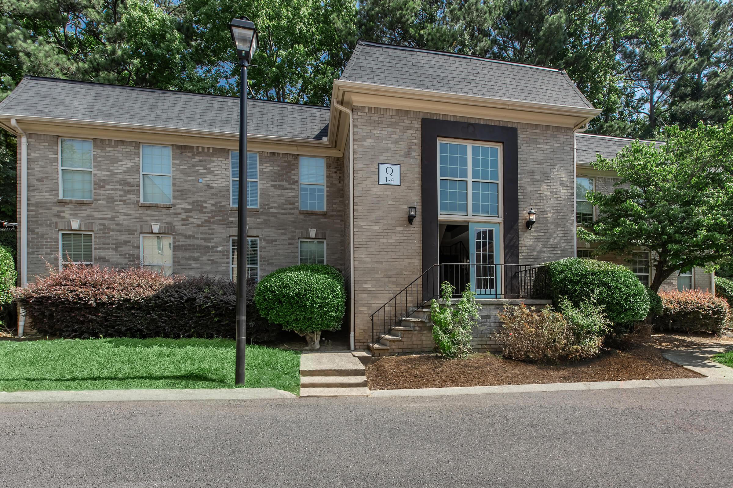 A two-story brick apartment building with a central entrance framed by large windows. The building has a neatly landscaped front yard featuring manicured shrubs and trees. A lamp post stands beside the sidewalk leading to the entrance. The scene is sunny, indicating a clear day.
