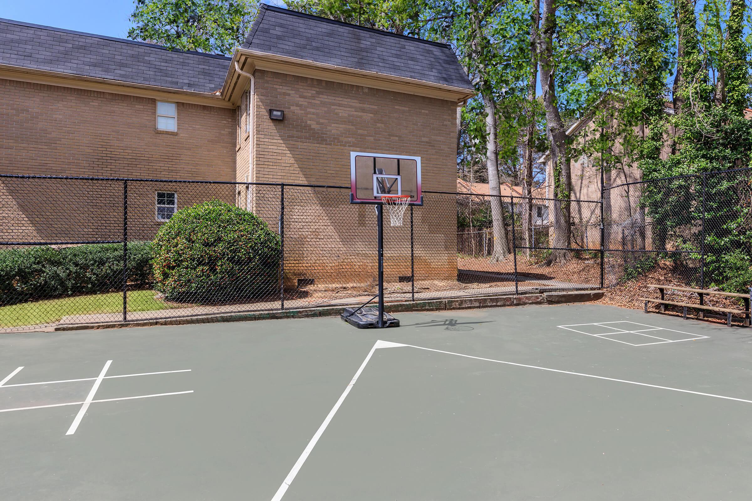 A basketball court featuring a hoop with a net, surrounded by a chain-link fence. The court has white markings for boundaries, and there are trees and a building in the background. The scene is bright and well-maintained, suggesting a recreational area for sports.