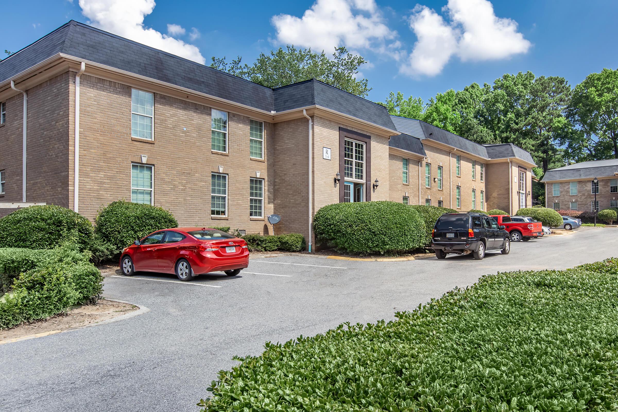 A brick apartment building with multiple windows surrounded by lush greenery. In the foreground, there are several parked cars, including a red sedan and a black SUV, along a paved driveway. The sky is clear with a few clouds, creating a bright and inviting atmosphere.