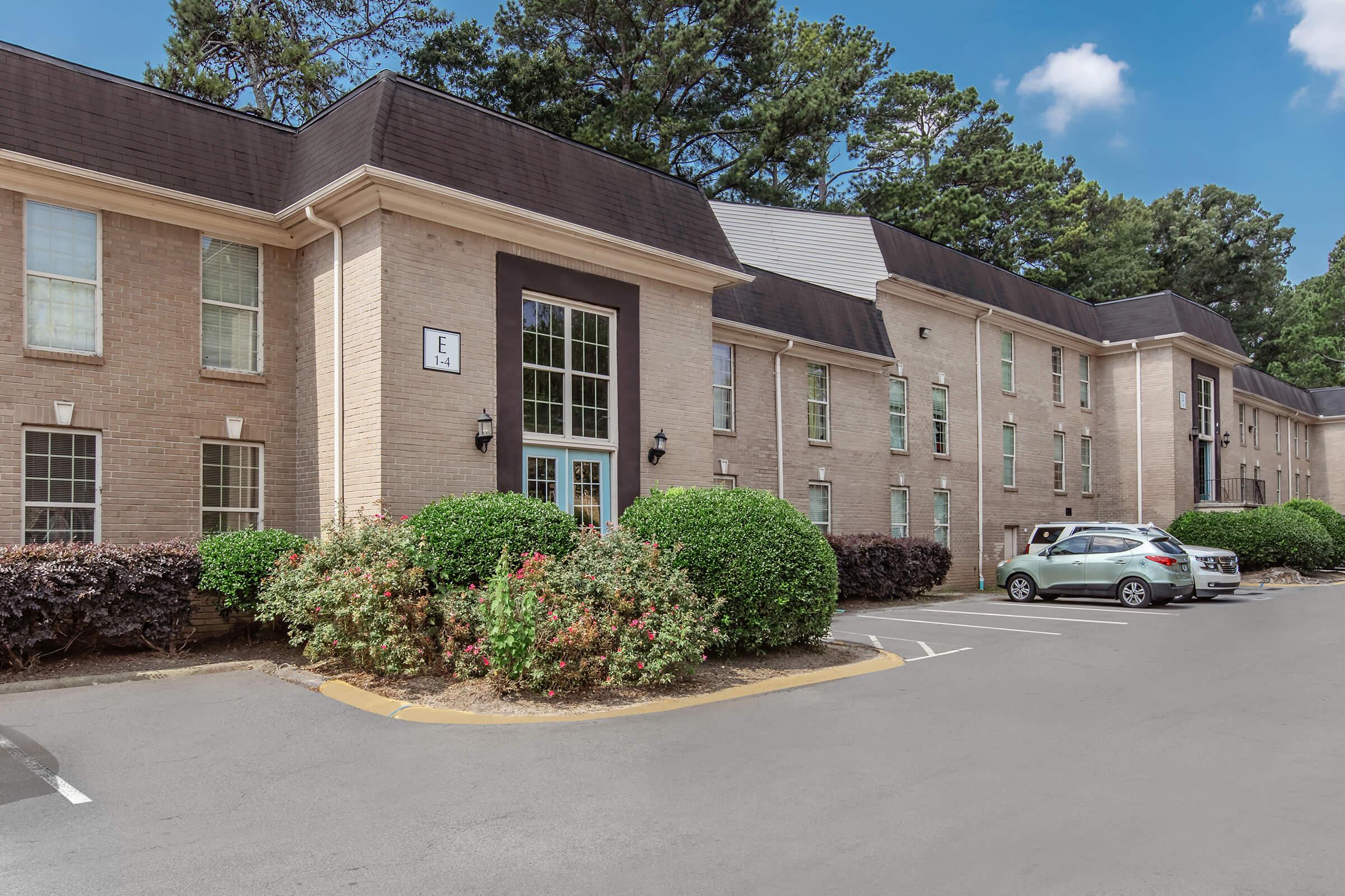 A beige multi-story building surrounded by well-maintained shrubs and trees. The entrance features large windows and is flanked by decorative lights. A parking area is visible in front with a parked white car. The sky is partly cloudy, creating a pleasant atmosphere.