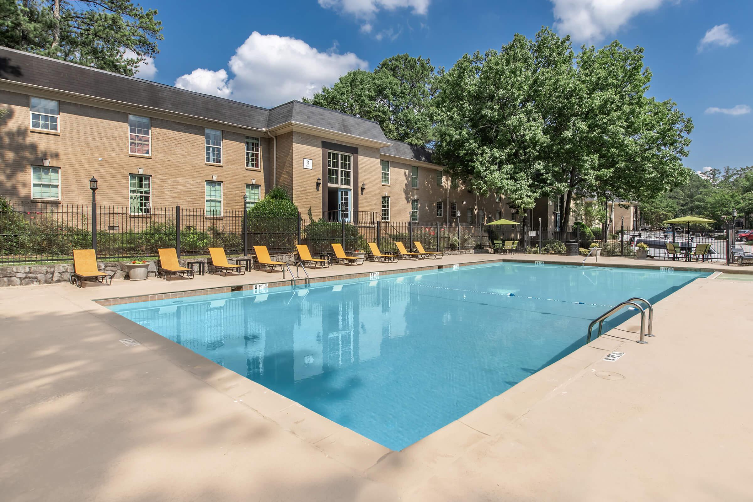 A clear swimming pool surrounded by lounge chairs and green trees. The pool area is part of a residential complex with a brick building in the background. The sky is partly cloudy, creating a bright and inviting atmosphere.