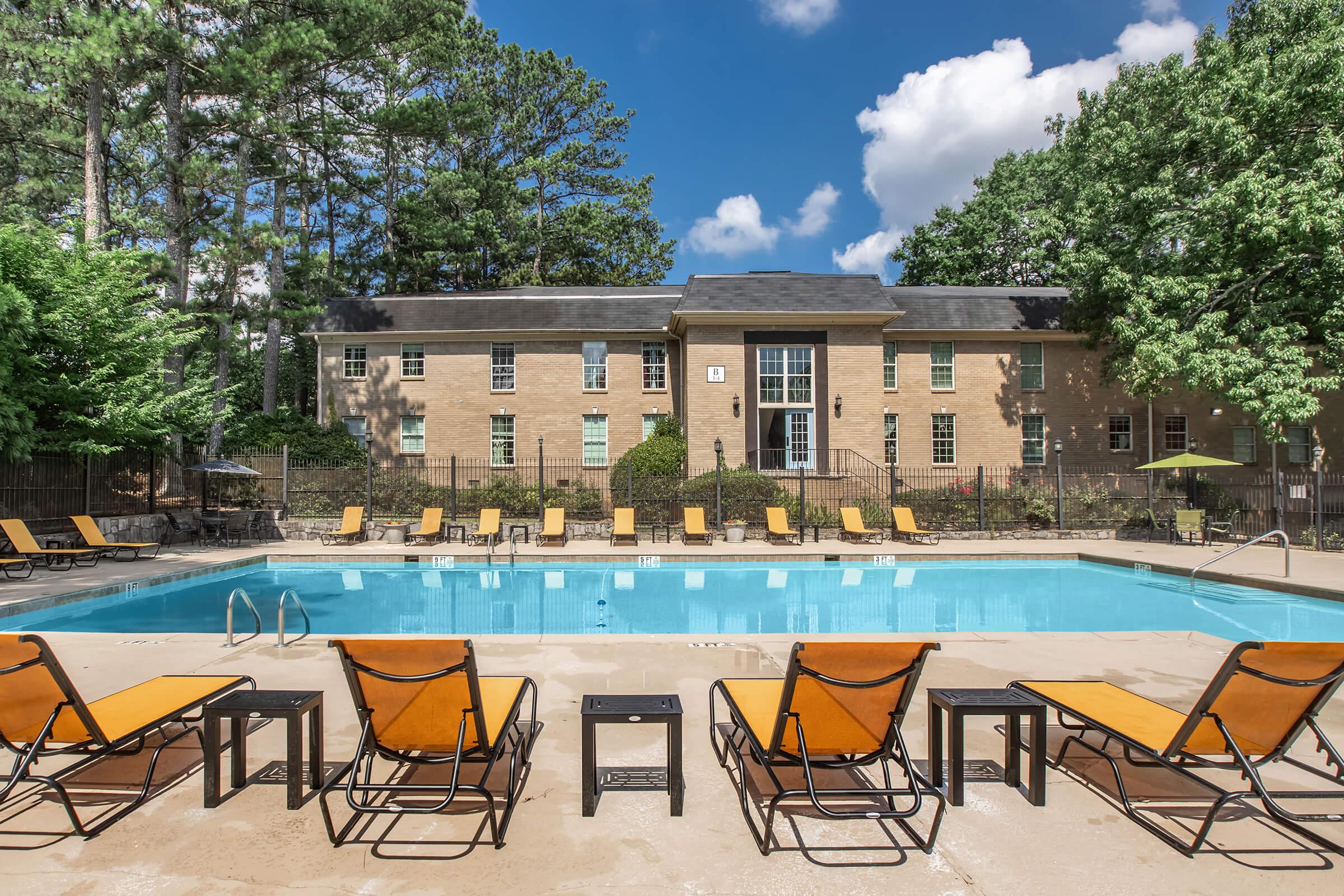 A sunny outdoor swimming pool surrounded by orange lounge chairs. A brick building with multiple windows is in the background, along with tall trees and a clear blue sky with fluffy clouds. The area appears inviting and well-maintained, perfect for relaxation and leisure.