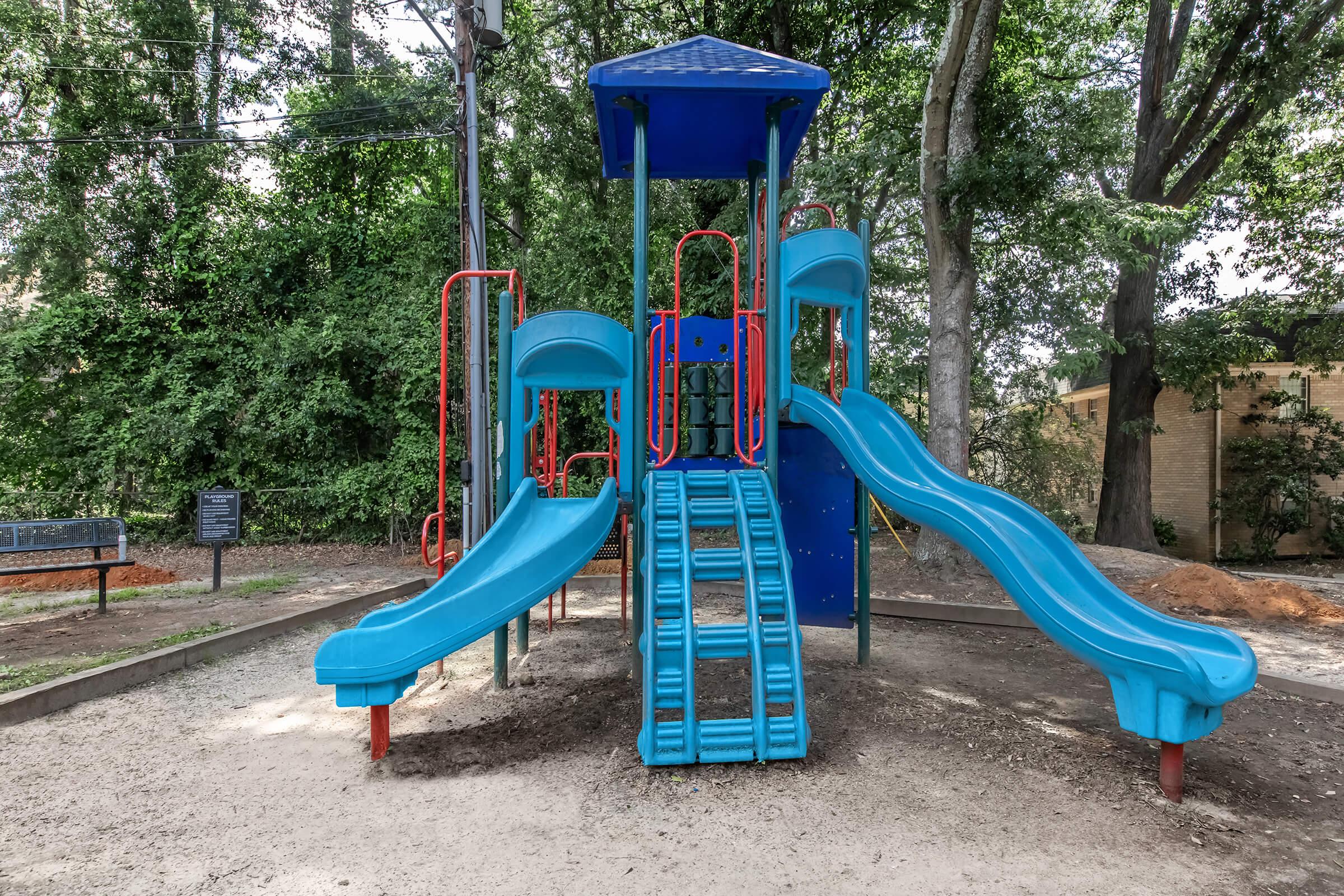 A colorful playground structure featuring two blue slides, a staircase for climbing, and a canopy. It is surrounded by greenery, with trees in the background and a bench visible nearby. The ground is covered in gravel, creating a safe play area for children.