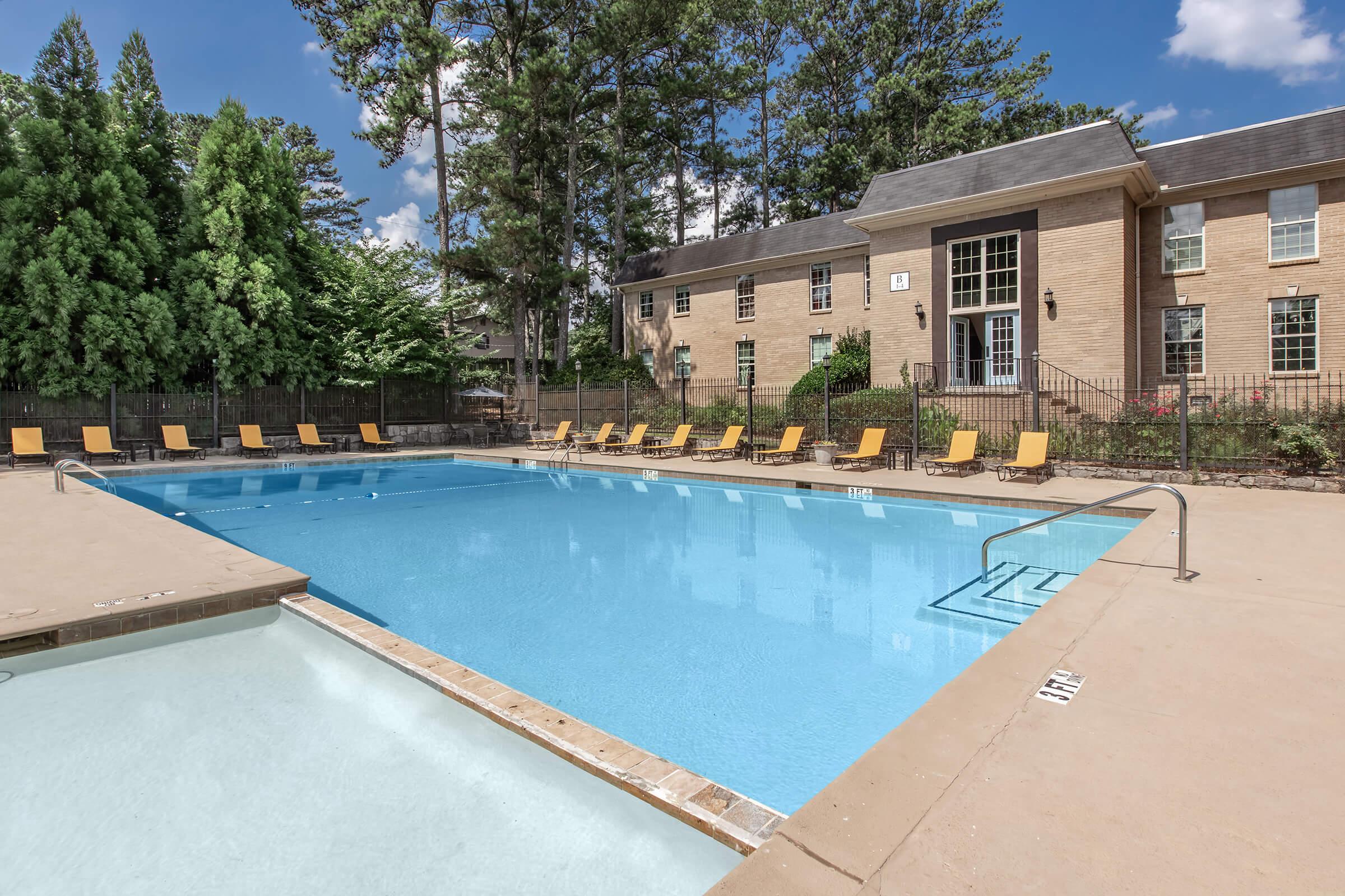 A clear swimming pool with a light blue surface, surrounded by several yellow lounge chairs. In the background, there is a tan brick building with large windows and a tree-lined area providing shade. The sky is bright blue with a few clouds, creating a welcoming outdoor atmosphere.