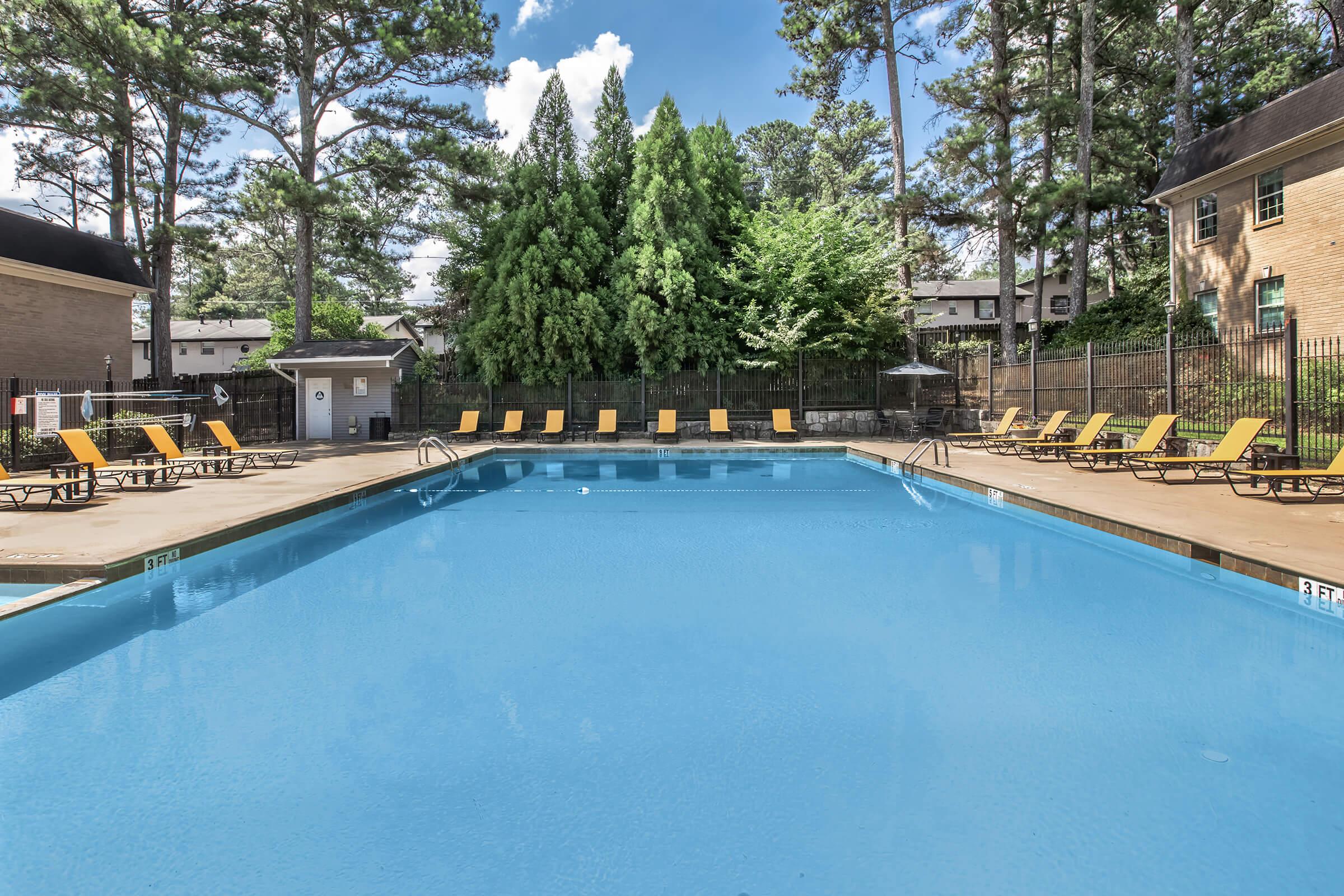 A well-maintained outdoor swimming pool surrounded by lounge chairs under a clear blue sky. Lush green trees are in the background, providing a tranquil atmosphere. The pool area appears clean and inviting, ideal for relaxation and recreation.