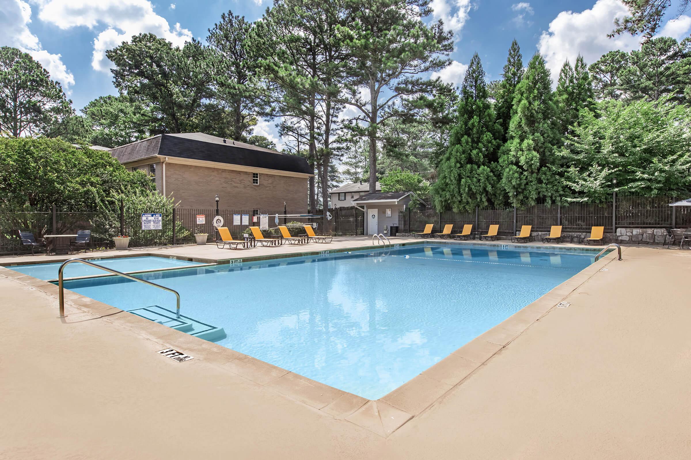 A clear swimming pool surrounded by a tan deck, with several yellow lounge chairs arranged around it. Lush green trees and shrubs are visible in the background, along with a two-story building. The sky is bright with fluffy white clouds.