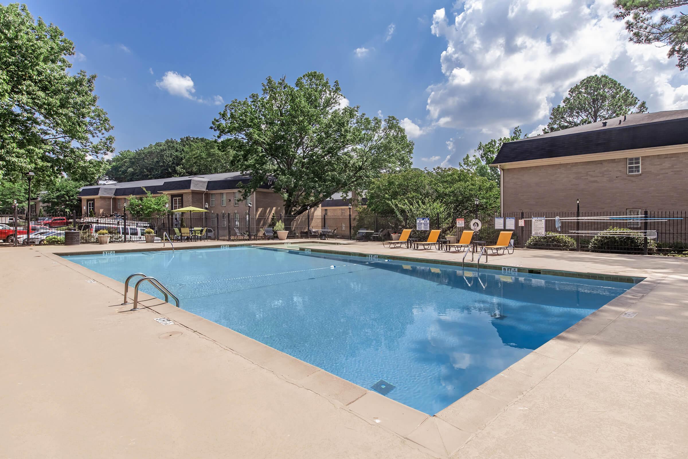A bright outdoor swimming pool surrounded by a concrete deck with lounge chairs. Lush green trees provide shade, and a clear blue sky with fluffy white clouds reflects in the water. In the background, several buildings are visible, creating a relaxing atmosphere for leisure and recreation.