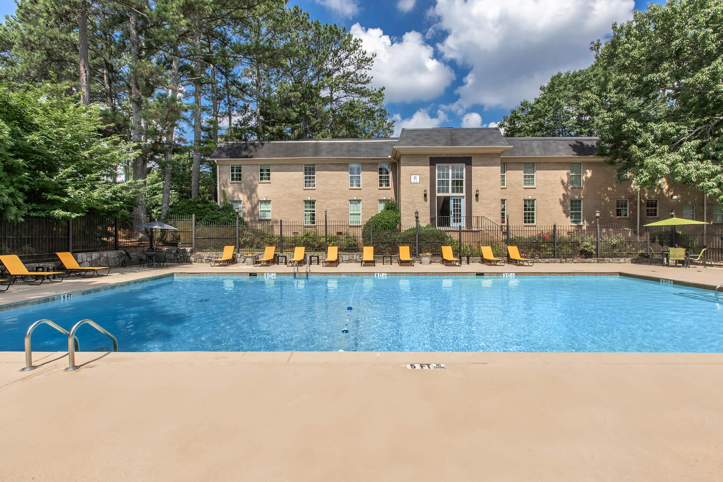 A clear blue swimming pool surrounded by lounge chairs with bright yellow cushions. In the background, there is a well-maintained brick building with multiple windows, set among trees under a partly cloudy sky. The pool area has a clean and inviting appearance, ideal for relaxation and leisure.