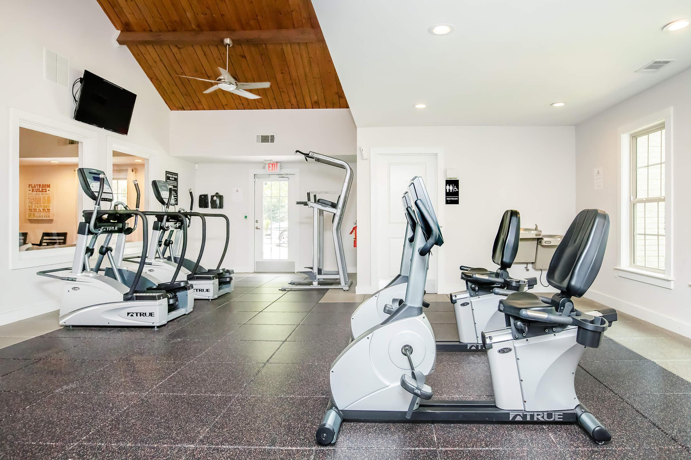 A modern gym interior featuring exercise equipment such as elliptical machines and recumbent bikes. The room has a high wooden ceiling, large windows for natural light, and rubber flooring. A flat-screen TV is mounted on one wall, creating a welcoming workout environment.