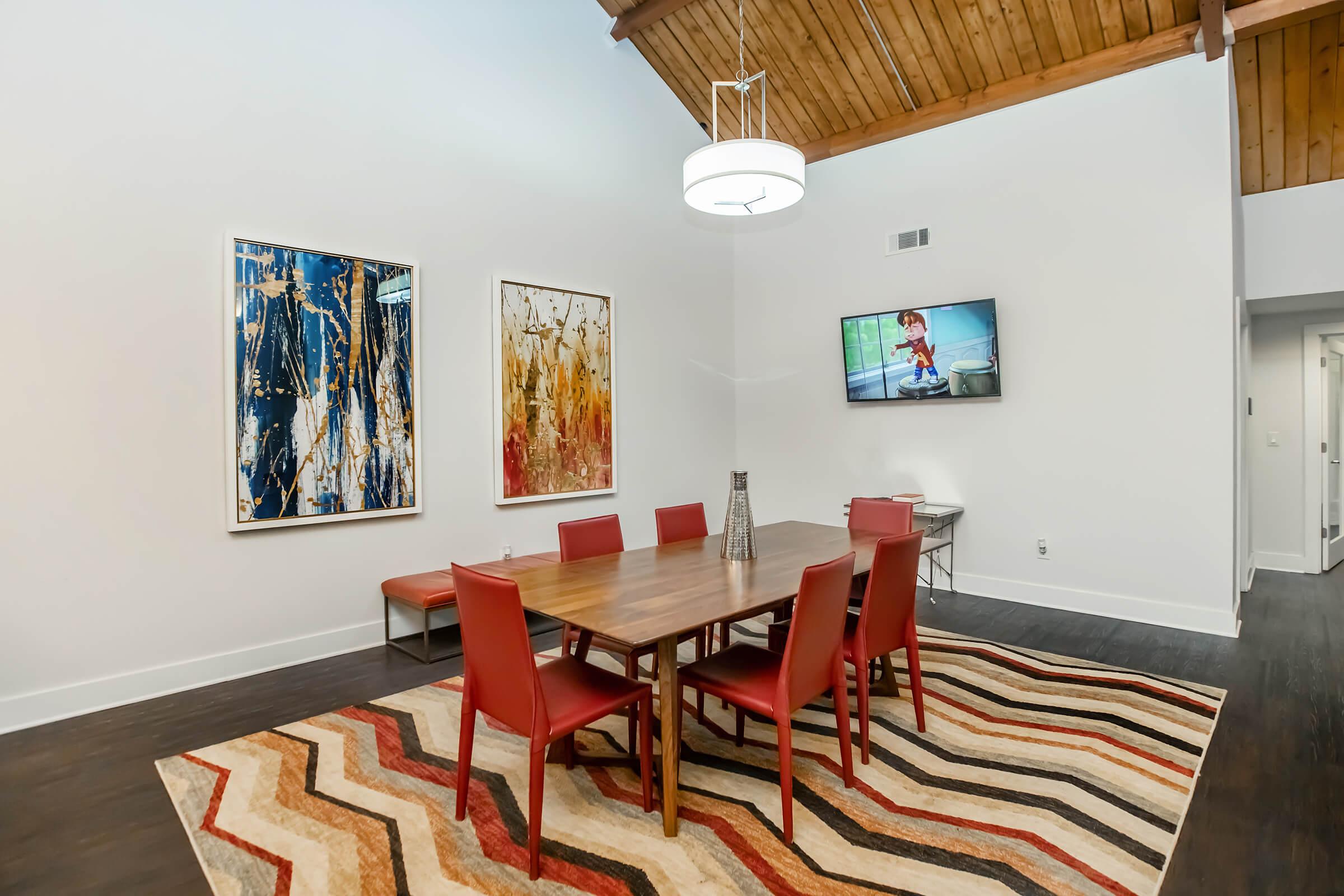A modern dining area featuring a wooden table surrounded by red chairs, a striped area rug, two large abstract paintings on the wall, and a mounted television displaying a program. The space has high ceilings with wooden beams and a contemporary light fixture.