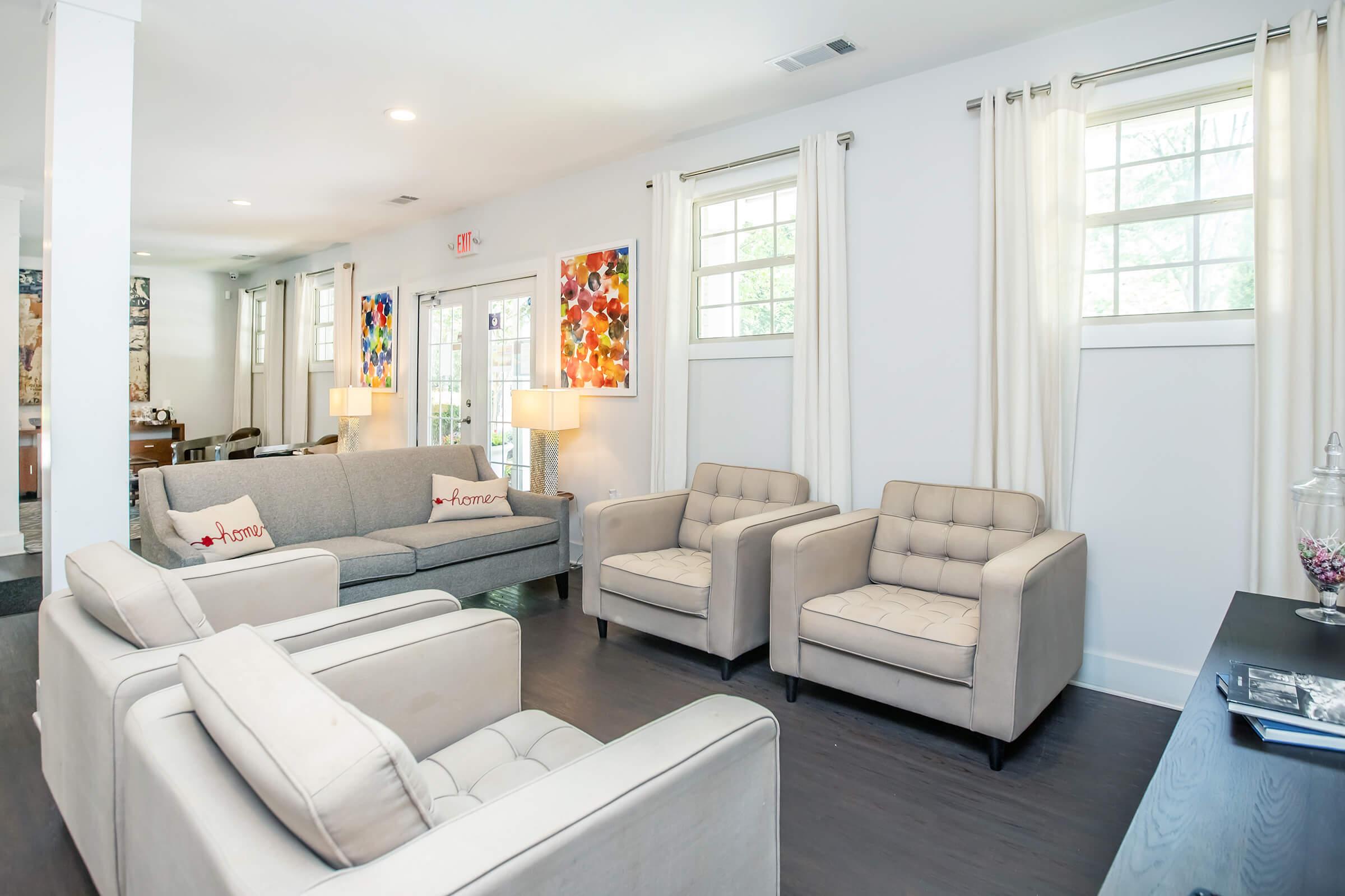A modern living room featuring a light gray sofa and two matching beige armchairs. The space has large windows with white curtains, two lamps providing warm lighting, and contemporary artwork on the walls. A dark wooden table is visible in the corner, adding to the elegant atmosphere.