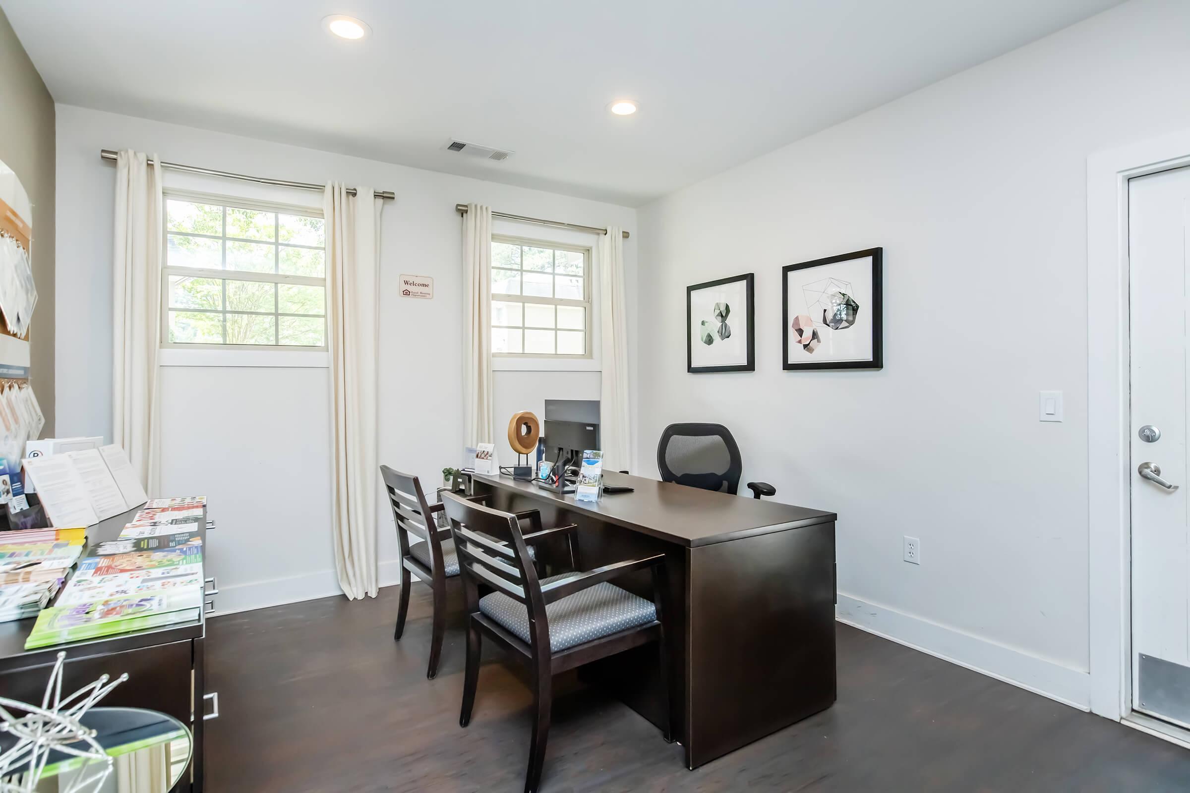 A modern office space featuring a wooden desk with a computer and an ergonomic chair, two framed artworks on the wall, large windows with sheer curtains, and a seating area with two chairs. A magazine rack and a door are visible, creating a clean and professional atmosphere.