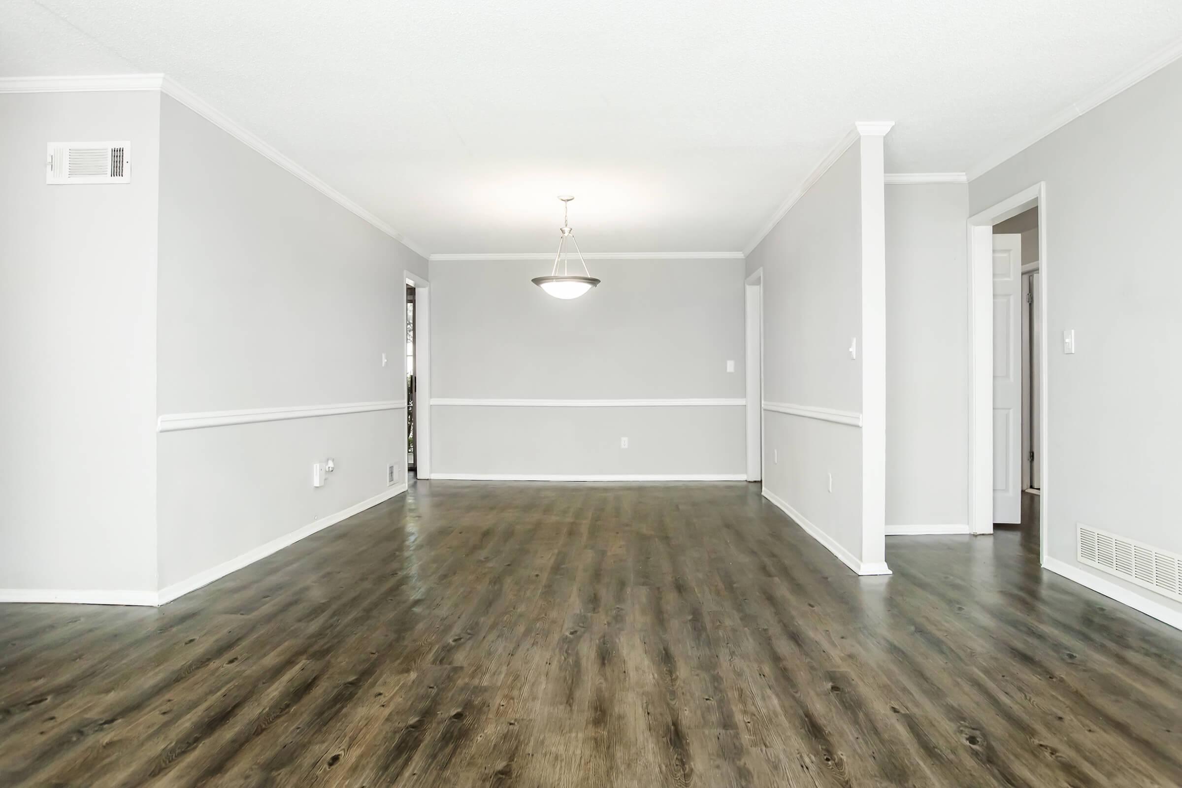 Interior view of a spacious, empty room with gray walls and a dark wooden floor. A pendant light hangs from the ceiling, and a chair rail runs along the walls. On the right, there are doors leading to other spaces, and the overall atmosphere is bright and open.