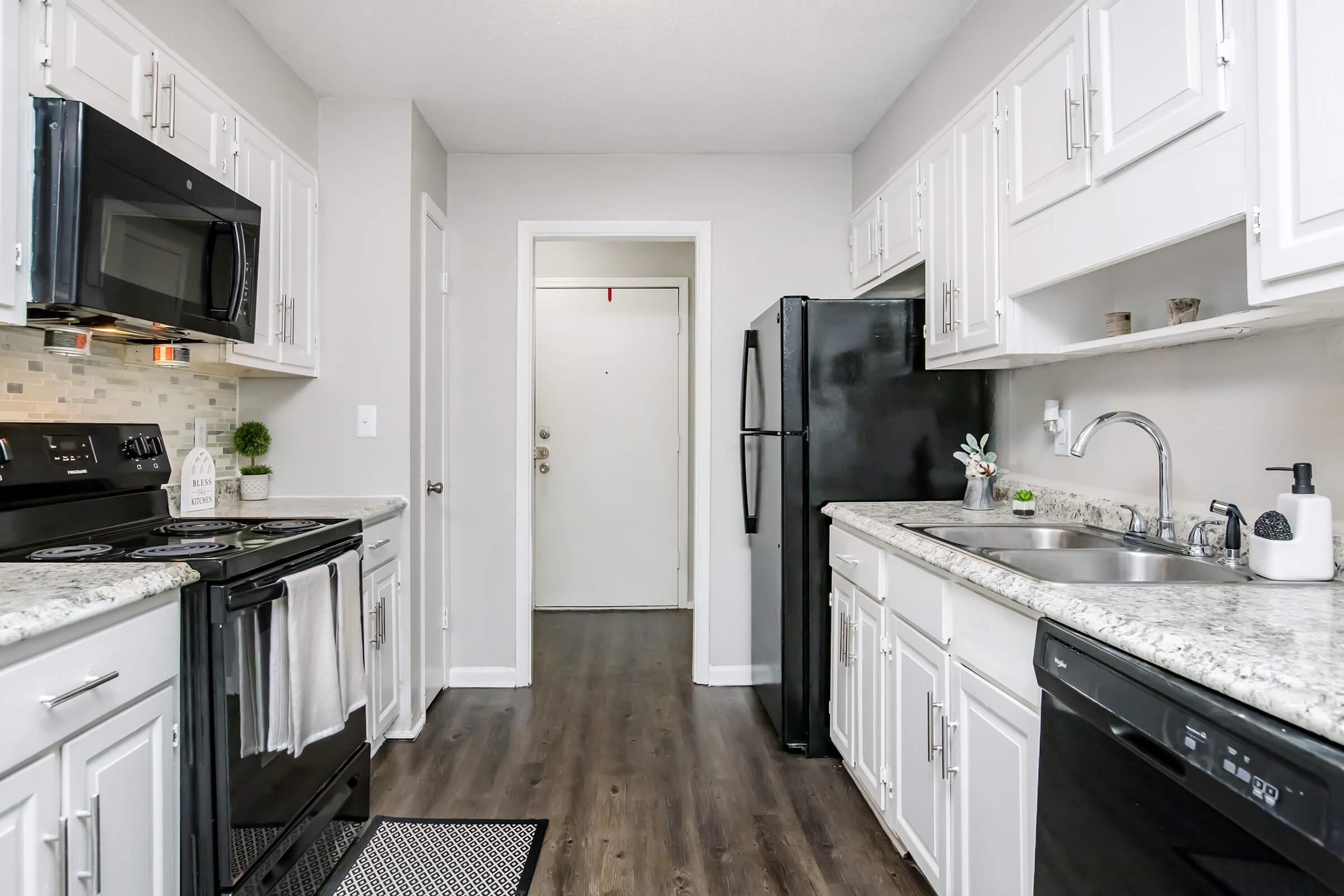 Modern kitchen featuring white cabinetry, black appliances, and a granite countertop. Includes a black refrigerator, oven, and dishwasher, with a sink under a window. Neutral-colored walls and wood-like flooring create a bright, clean atmosphere. A front door is visible in the background.