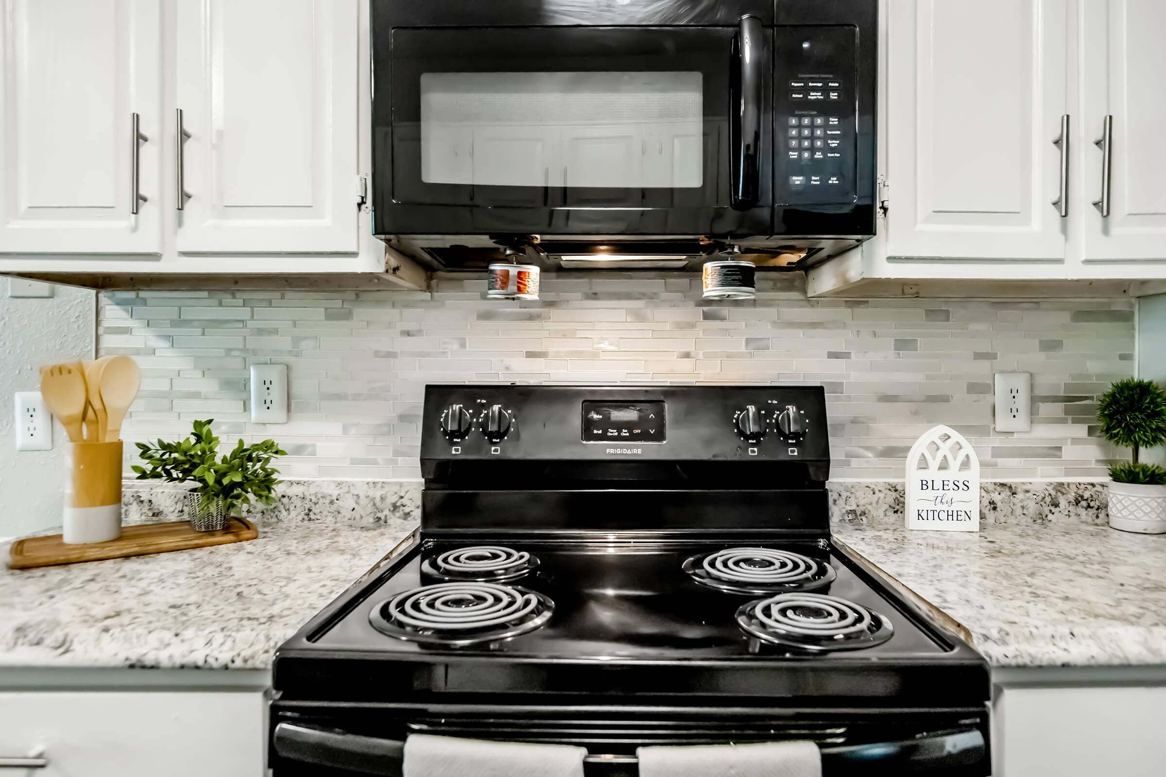 A modern kitchen featuring a black stove and microwave above, with a granite countertop. There are wooden utensils in a holder, small green plants for decor, and a decorative sign that reads "Bless this kitchen." The backsplash consists of light-colored tiles.