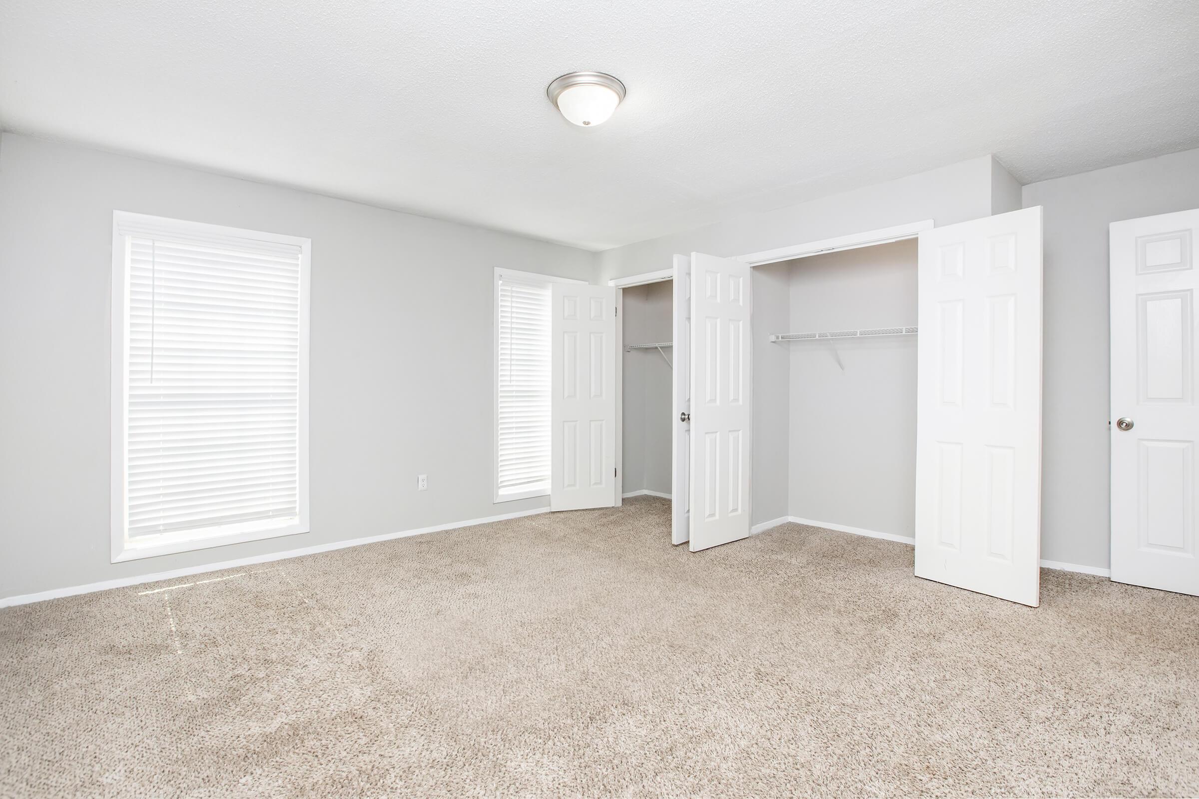 A spacious, empty room with light gray walls and beige carpet. Two large windows with white blinds let in natural light. There are two white closet doors opened, revealing a shelf inside. The ceiling has a simple light fixture. The overall atmosphere is bright and airy, ideal for personalized decoration.