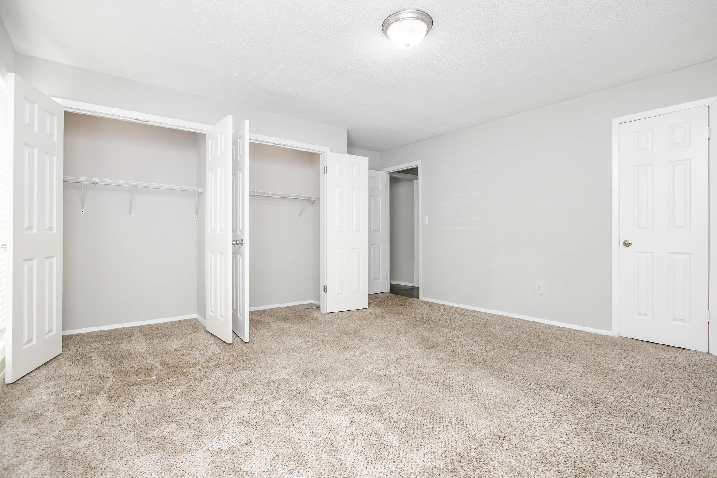 A spacious, empty room with light gray walls and beige carpet. Two white doors are slightly open, revealing closets with hanging rods. A door on the right leads to another area. The room is bright, featuring a ceiling light fixture.