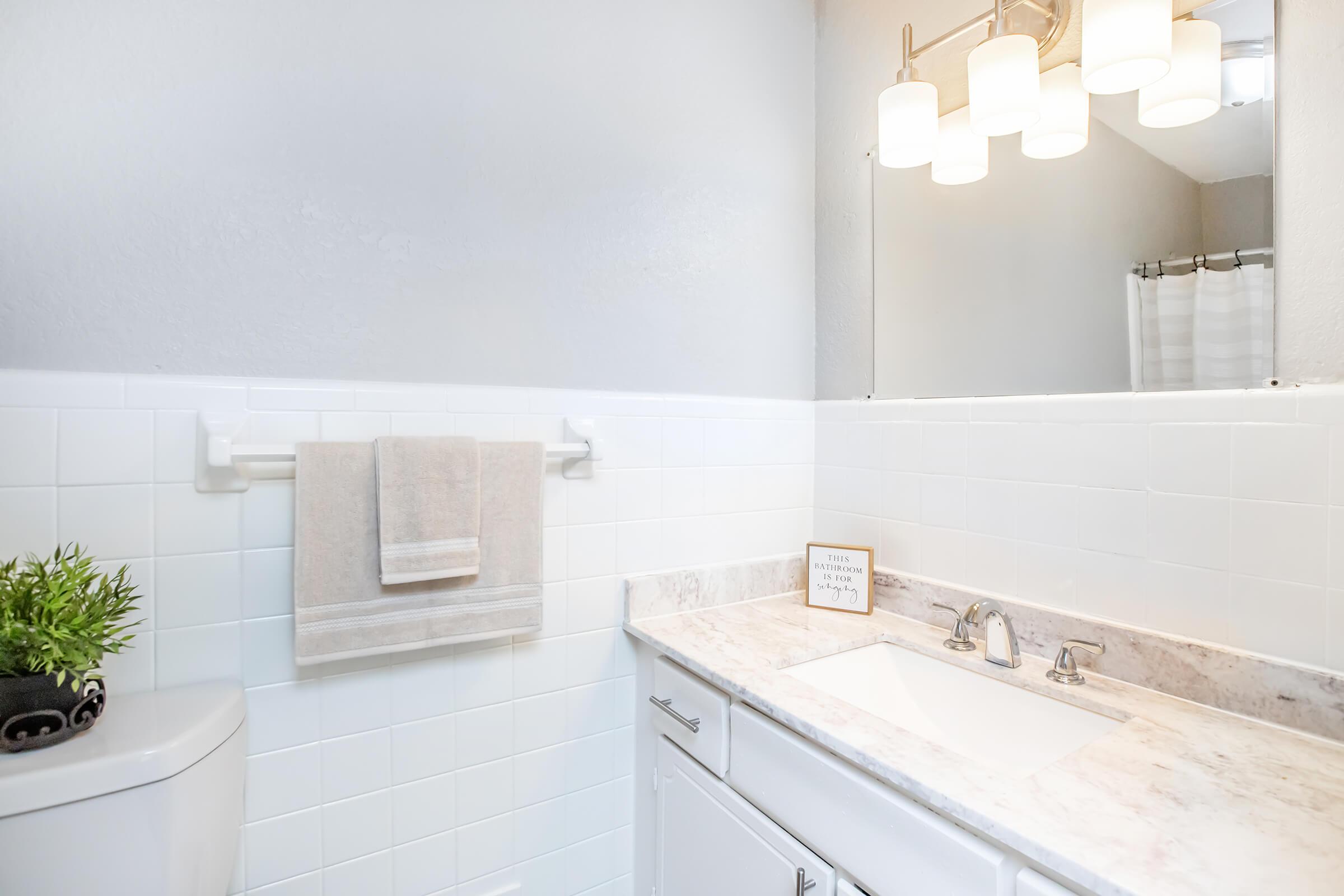 A clean bathroom featuring a white tiled wall, a modern sink with a marble countertop, and a towel on a rack. There's a small plant on the counter, and a mirror above the sink, illuminated by stylish light fixtures. A shower curtain is visible in the background.