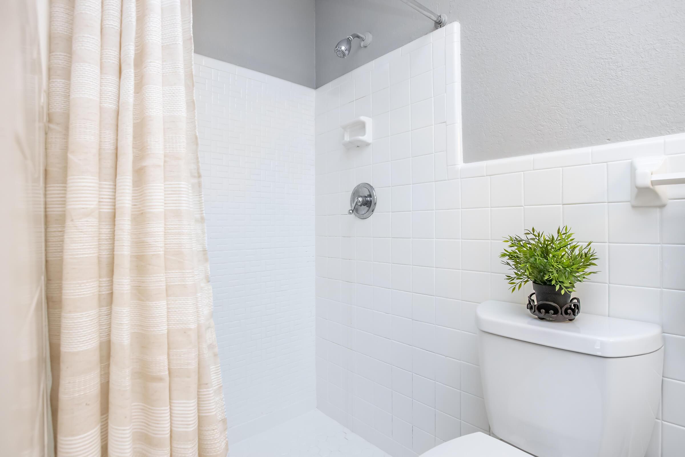 A clean bathroom featuring a shower area with a tiled wall, a showerhead, and a textured shower curtain. A small potted plant sits on the ledge, while a white toilet is positioned nearby, emphasizing a bright and minimalist design.