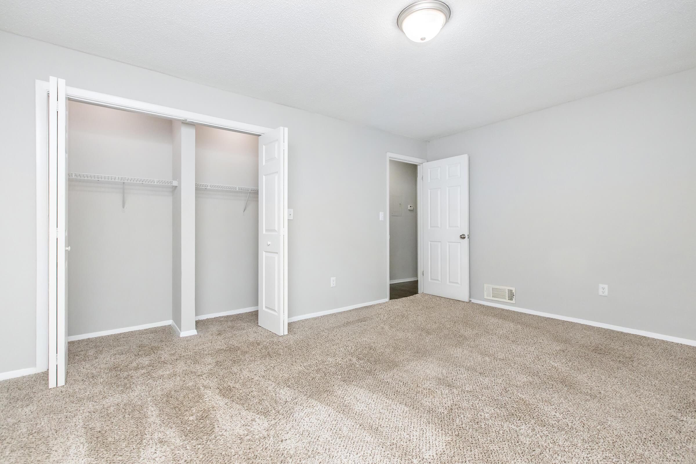 A clean, well-lit bedroom featuring tan carpet flooring and light gray walls. There are two open white doors leading to a closet with shelving and another door visible on the right, suggesting access to a bathroom or hallway. The ceiling is adorned with a circular light fixture.