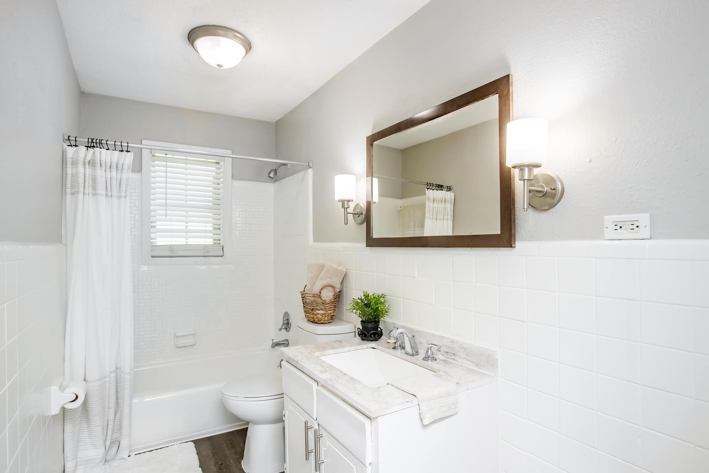 A modern bathroom featuring a white bathtub with a shower curtain, a wall-mounted mirror with a light fixture, a vanity with a marble countertop, and a decorative plant. The walls are painted light gray, and there is a small window with blinds, allowing natural light to enter the space.