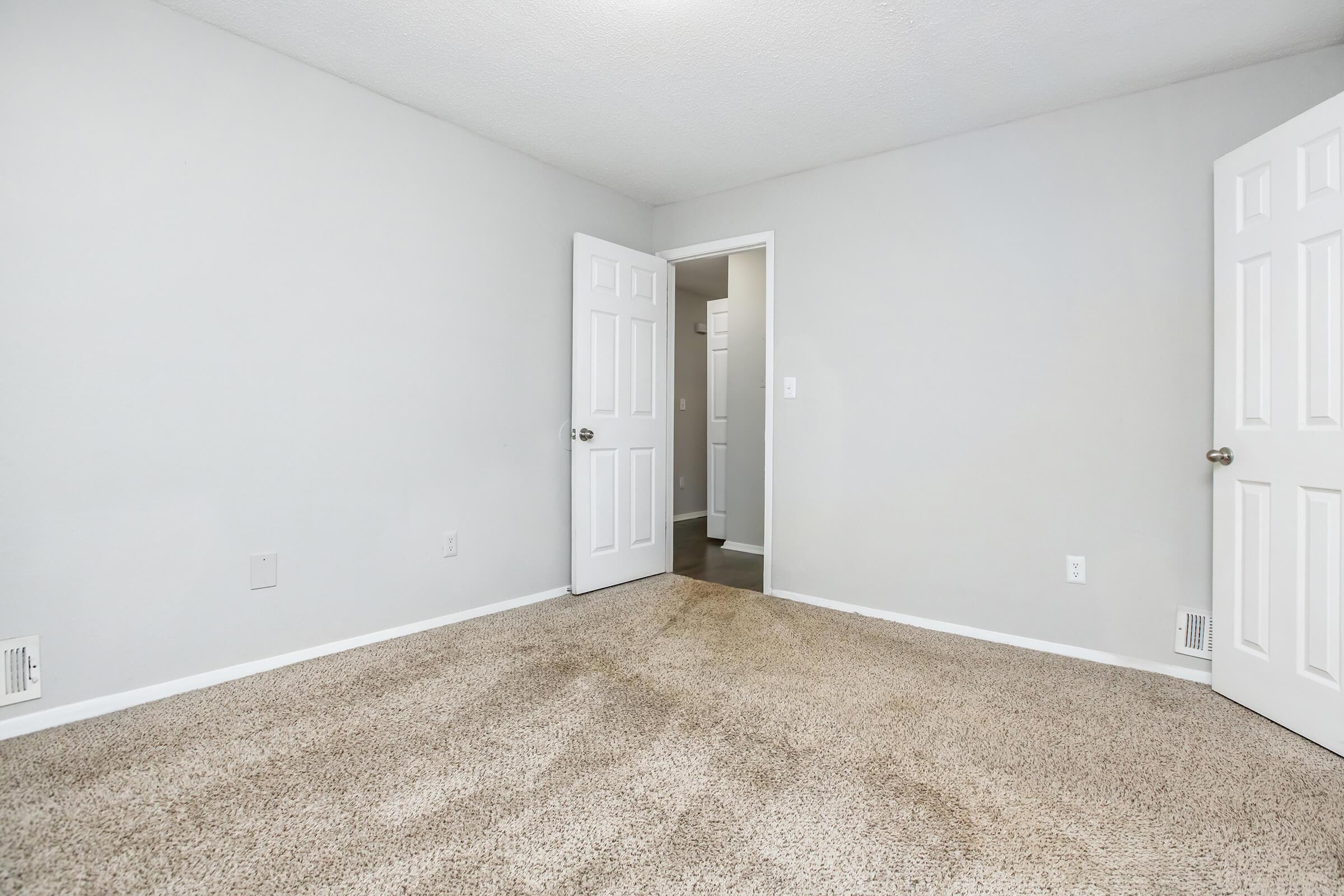 Empty room with light gray walls and beige carpet. There are two white doors, one leading to a small hallway and the other opening outward. The space is well-lit, creating a neutral and airy atmosphere, suitable for various purposes such as a bedroom or office.