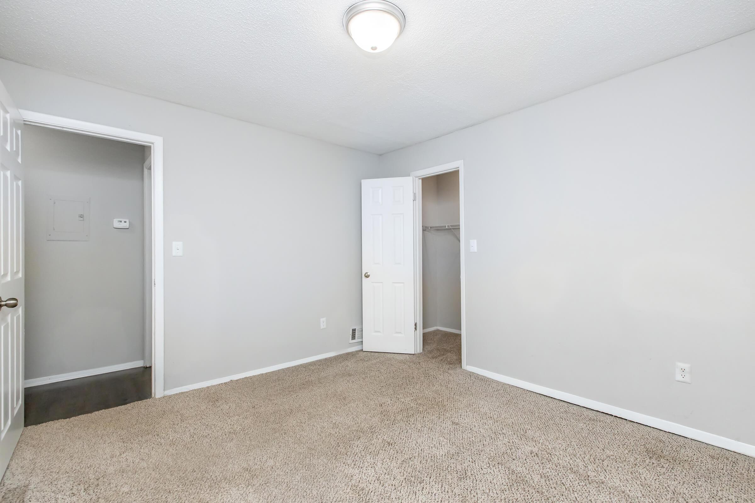 A light-filled room with beige carpet flooring and light gray walls. It features two doorways: one leading to a closet and another exiting to a hallway. The ceiling has a simple light fixture, creating a clean and spacious atmosphere, ideal for various uses such as a bedroom or office.