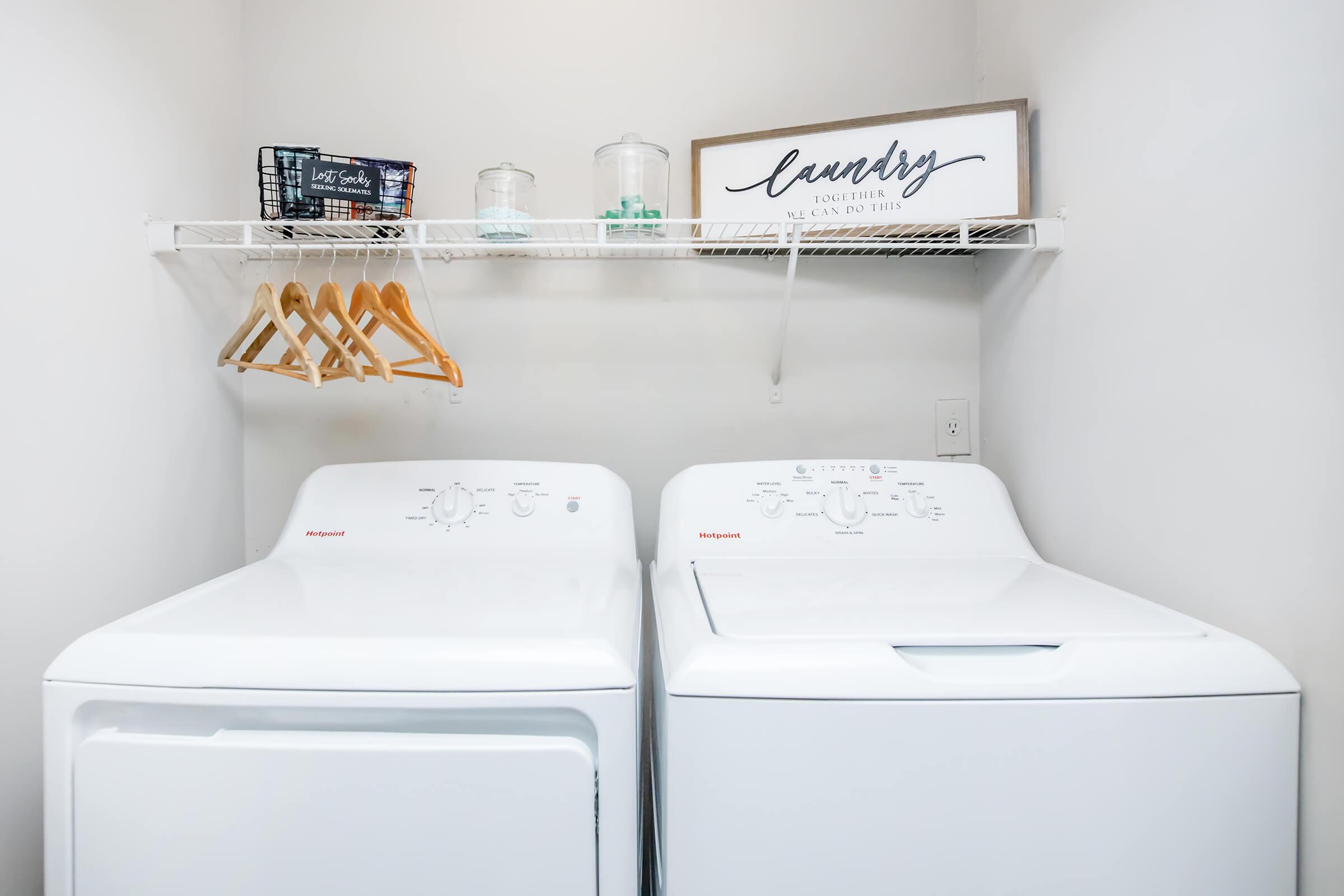 A clean laundry room featuring a stacked washer and dryer, with a shelf above holding glass containers and a decorative sign that reads "laundry." There are wooden hangers on the shelf, and the walls are painted in a light color to create a bright, airy atmosphere.