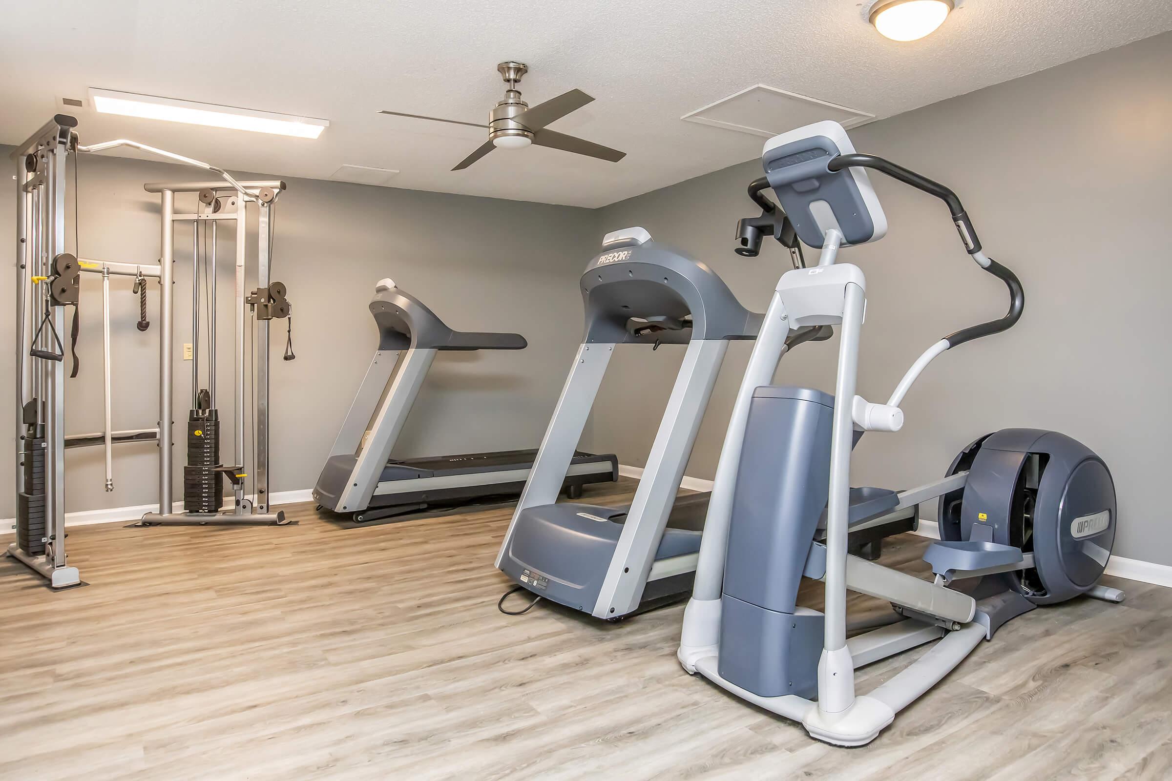 A modern gym interior featuring two treadmills, an elliptical machine, and a multi-gym strength training machine. The room has gray walls, wooden flooring, and a ceiling fan, creating a bright and inviting workout space.