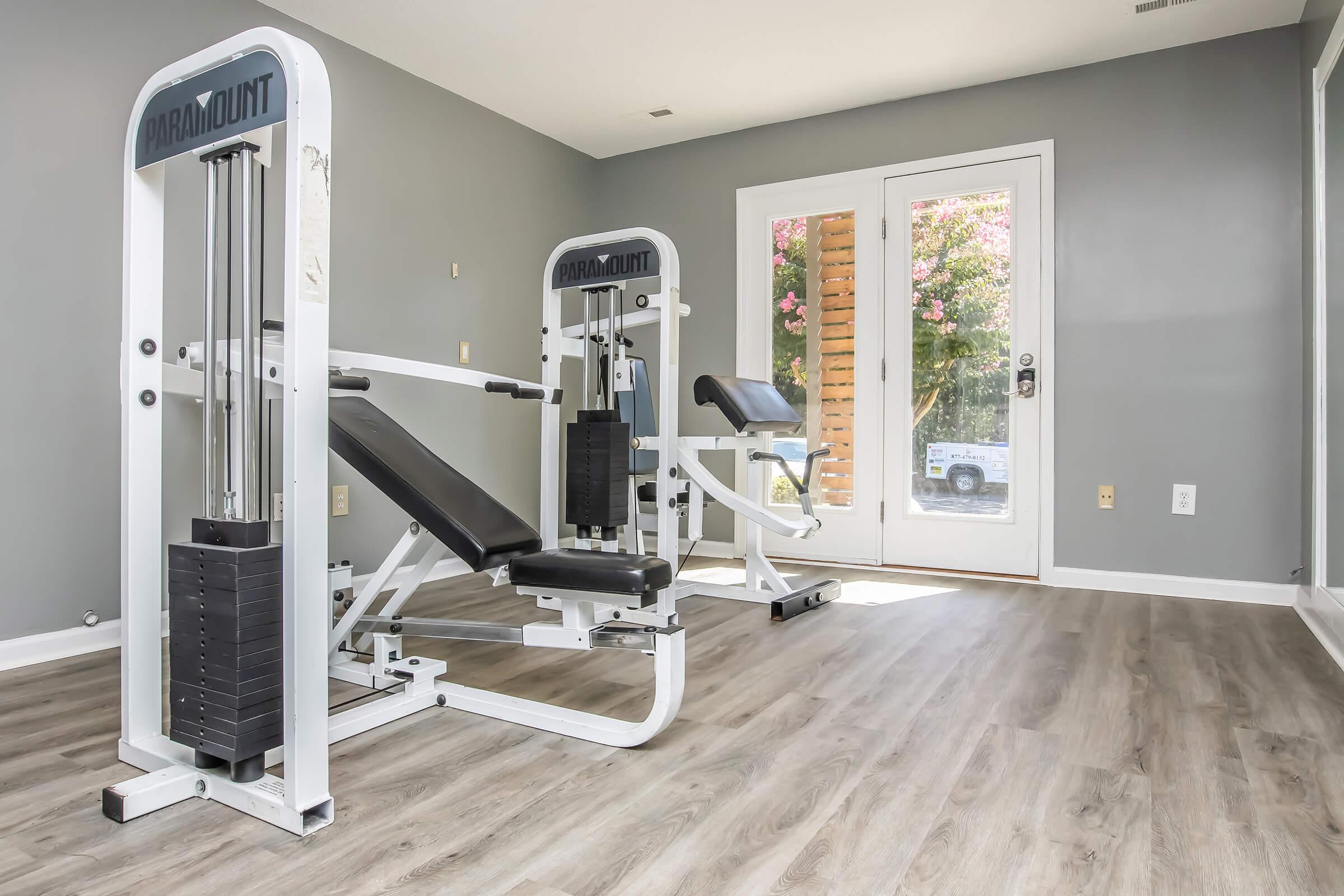 A well-lit home gym featuring a multi-station workout machine and a weight bench. The room has gray walls and large windows allowing natural light, with a view of greenery outside. The flooring is a light-colored wood laminate, creating a modern and inviting exercise space.
