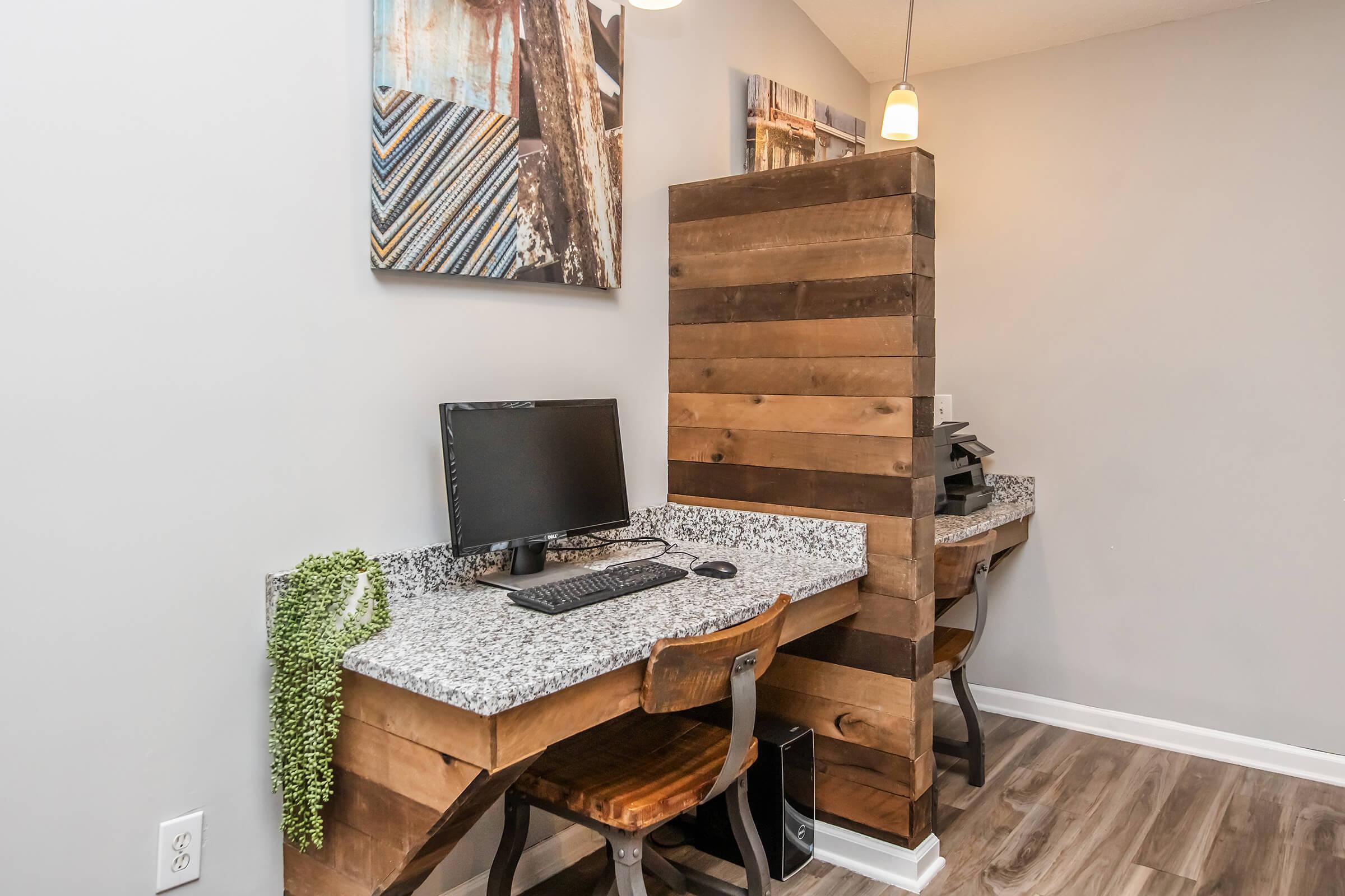 A cozy workspace featuring two wooden desks with granite surfaces, each equipped with a computer and a comfortable chair. A wooden partition separates the desks, and decorative wall art adds a touch of style. The space is well-lit with a pendant light and has a modern, inviting atmosphere.